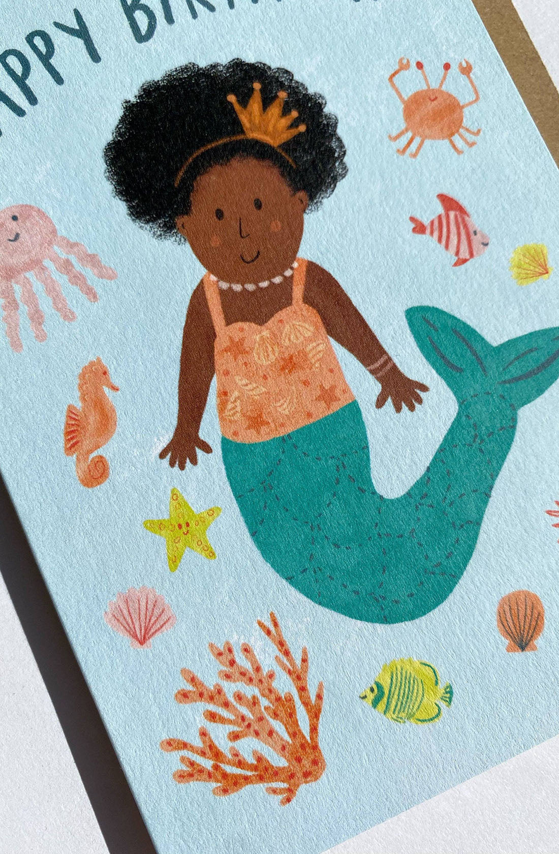 The Birthday Greeting Card Mermaid "Happy Birthday" by LAUREN SISSONS STUDIO features an illustrated dark-skinned mermaid with curly hair, an orange top, and a turquoise tail. Adorned with a crown and necklace, she is surrounded by colorful sea creatures including a jellyfish, starfish, seahorse, crab, and fish. This charming card is printed on recycled paper.