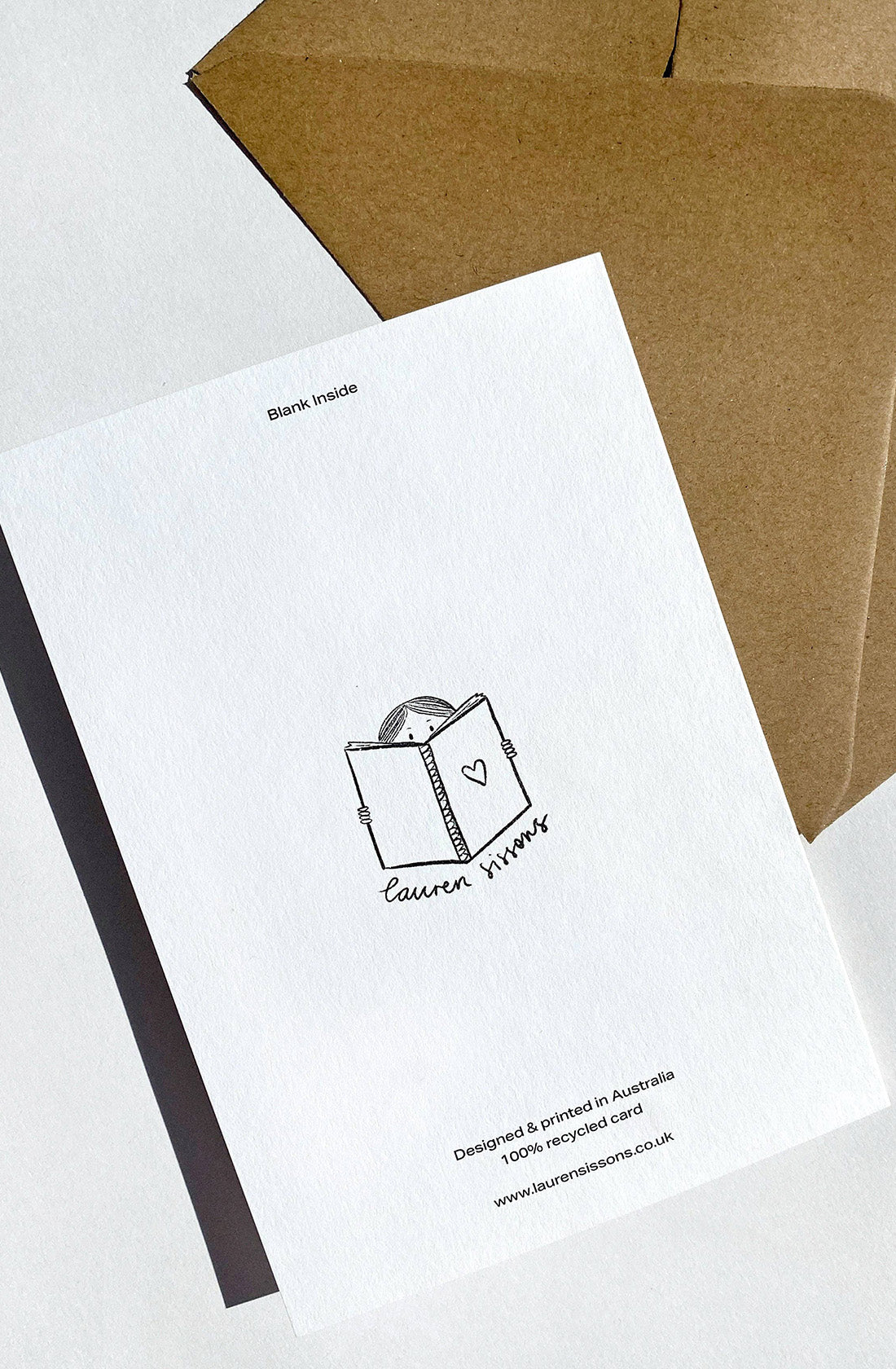 A birthday greeting card featuring a simple illustration of a book with a heart on the cover and a face peeking over it. The card, named "Happy Birthday" from LAUREN SISSONS STUDIO, is labeled "Blank Inside" and is placed on a brown envelope. Text at the bottom states that the card is printed on 100% recycled paper and includes a website.