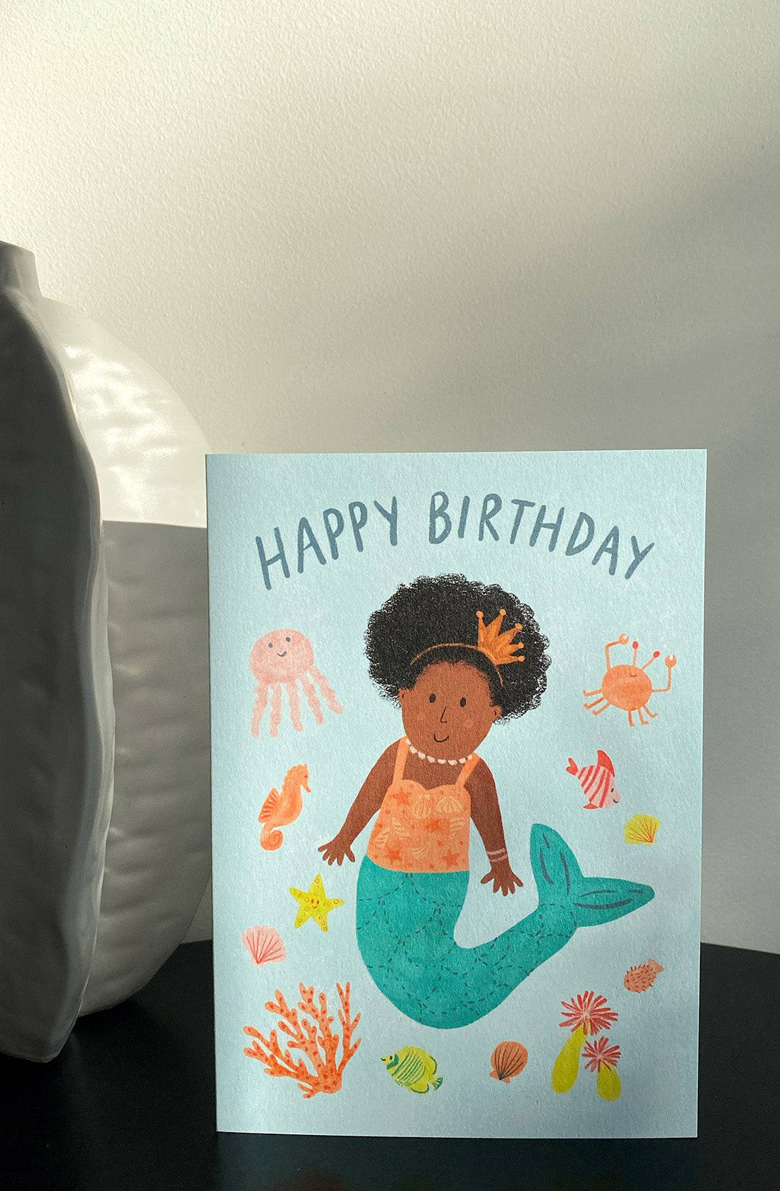 The LAUREN SISSONS STUDIO "Birthday Greeting Card Mermaid 'Happy Birthday'" features a vibrant mermaid theme, showcasing a cheerful young mermaid with curly hair and a crown. The background is adorned with illustrations of sea creatures such as a jellyfish, crab, seahorse, and corals. The text "HAPPY BIRTHDAY" is prominently displayed above her. Crafted from recycled paper, the card is displayed propped against a white vase.