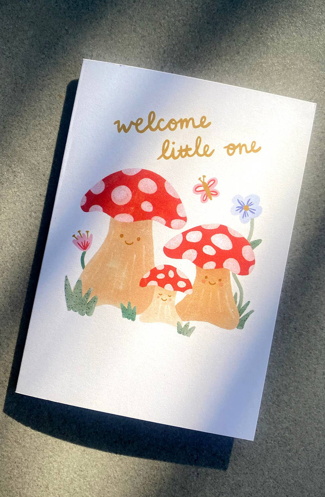 The "New Baby Greeting Card 'Welcome Little One'" from LAUREN SISSONS STUDIO is a gender-neutral card featuring three adorable cartoon mushrooms with red caps and white spots, accompanied by two flowers and lush green grass. The gold cursive text "welcome little one" elegantly floats above the mushrooms. Made from recycled paper, this charming card is showcased on a gray surface that casts gentle shadows.