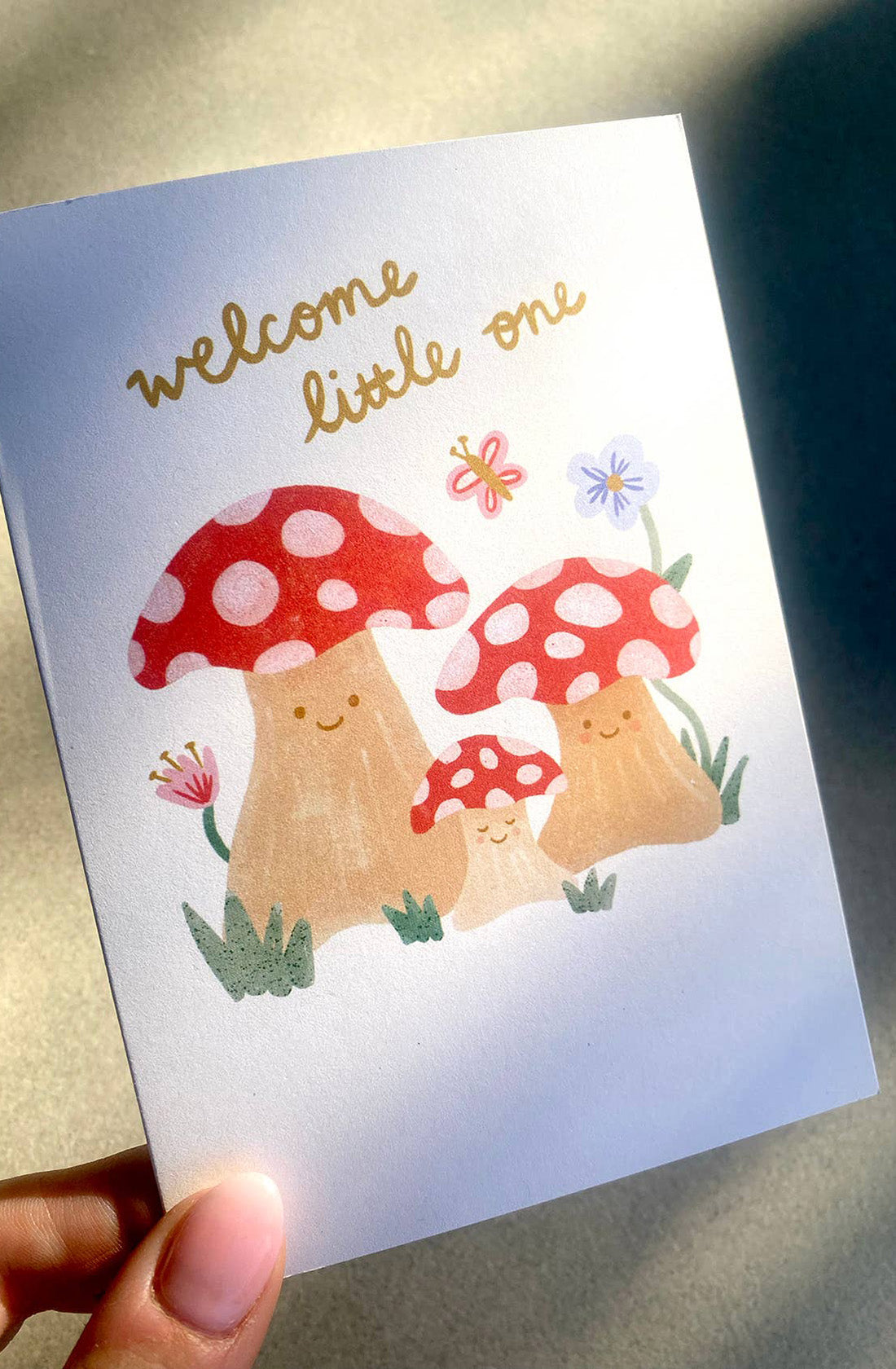 Introducing the New Baby Greeting Card "Welcome Little One" Mushrooms by LAUREN SISSONS STUDIO. This gender-neutral card, crafted from recycled paper, features an adorable illustration of three smiling mushrooms with red and white polka-dotted caps. Above them, shimmering in gold cursive, reads "Welcome little one." Small colorful flowers and butterflies surround the cheerful mushrooms, making it a perfect way to celebrate a new arrival.