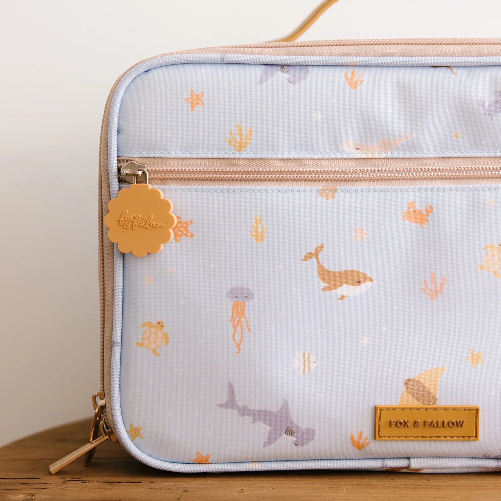 The Ocean Creatures Sky Lunch Bag by FOX & FALLOW is a light blue, kid-approved insulated lunch bag featuring a peach zipper and handle. It is adorned with marine life illustrations including a whale, jellyfish, and shark. A small gold zipper charm has "Fox & Fallow" engraved, and the brand name "FOX & FALLOW" appears on a leather patch.