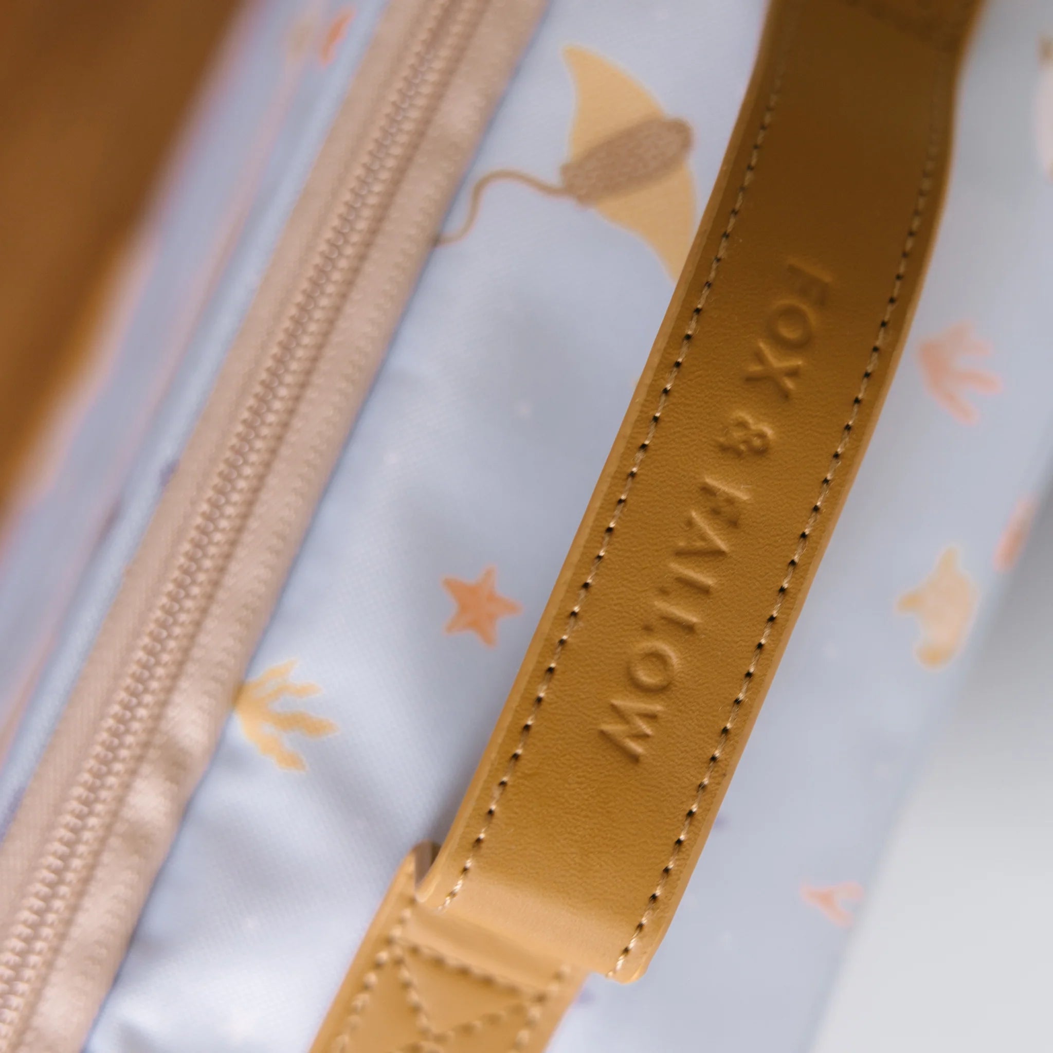 Close-up of a leather strap on the Ocean Creatures Sky Lunch Bag by FOX & FALLOW, showcasing the embossed text "FOX & FALLOW." The lunch bag features a light blue background adorned with whimsical patterns of starfish, manta rays, and other ocean-themed illustrations. Made from waterproof fabric, it's perfect for kid-approved adventures.