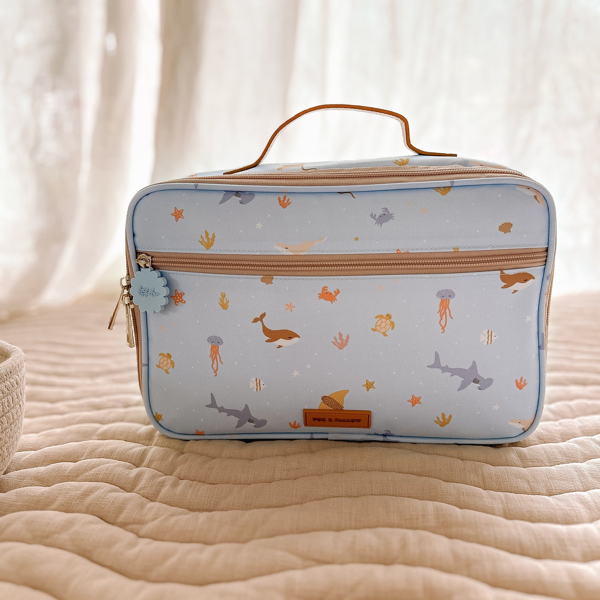 The Ocean Creatures Sky Lunch Bag by FOX & FALLOW is a light blue children's backpack with a brown handle. It features delightful sea-themed illustrations of whales, sharks, crabs, jellyfish, and starfish. Made from waterproof fabric, the bag is displayed on a beige quilted surface and includes a front zipper pocket ideal for insulated lunch bags.