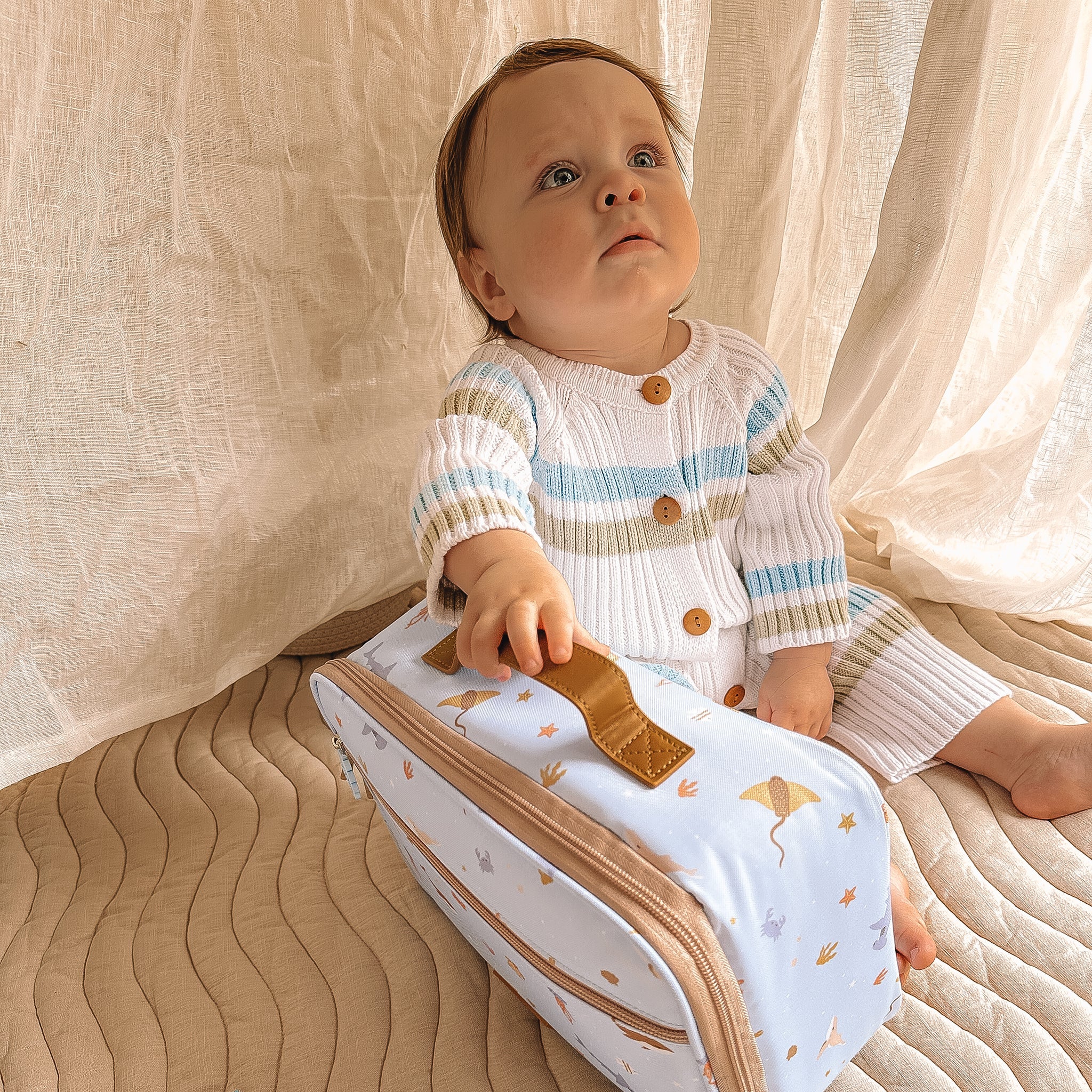 A baby with light hair and a striped sweater sits on a quilted surface, holding the **FOX & FALLOW Ocean Creatures Sky Lunch Bag** featuring kite patterns and tan handles. The kid-approved lunch bag, made from waterproof fabric, captures the baby's curious gaze. A beige curtain hangs behind them.