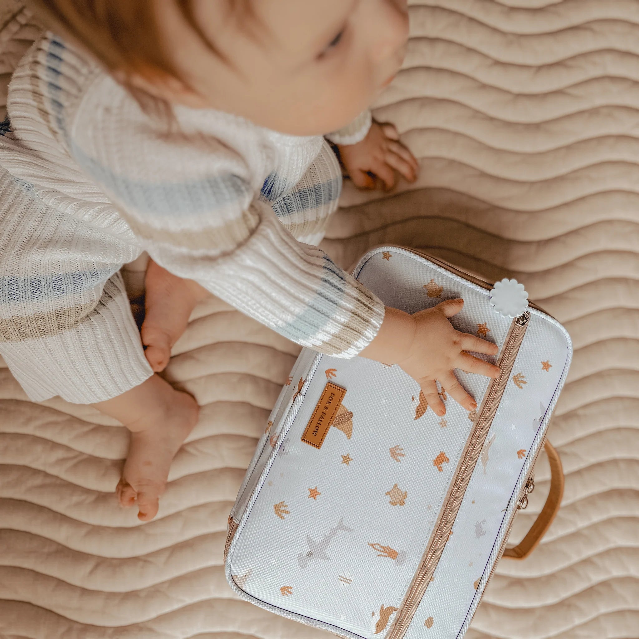 A baby wearing a striped sweater and ribbed pants sits on a quilted surface, reaching out to the Ocean Creatures Sky Lunch Bag by FOX & FALLOW, made from waterproof fabric and featuring an animal print with foxes, bears, and birds on a white background.