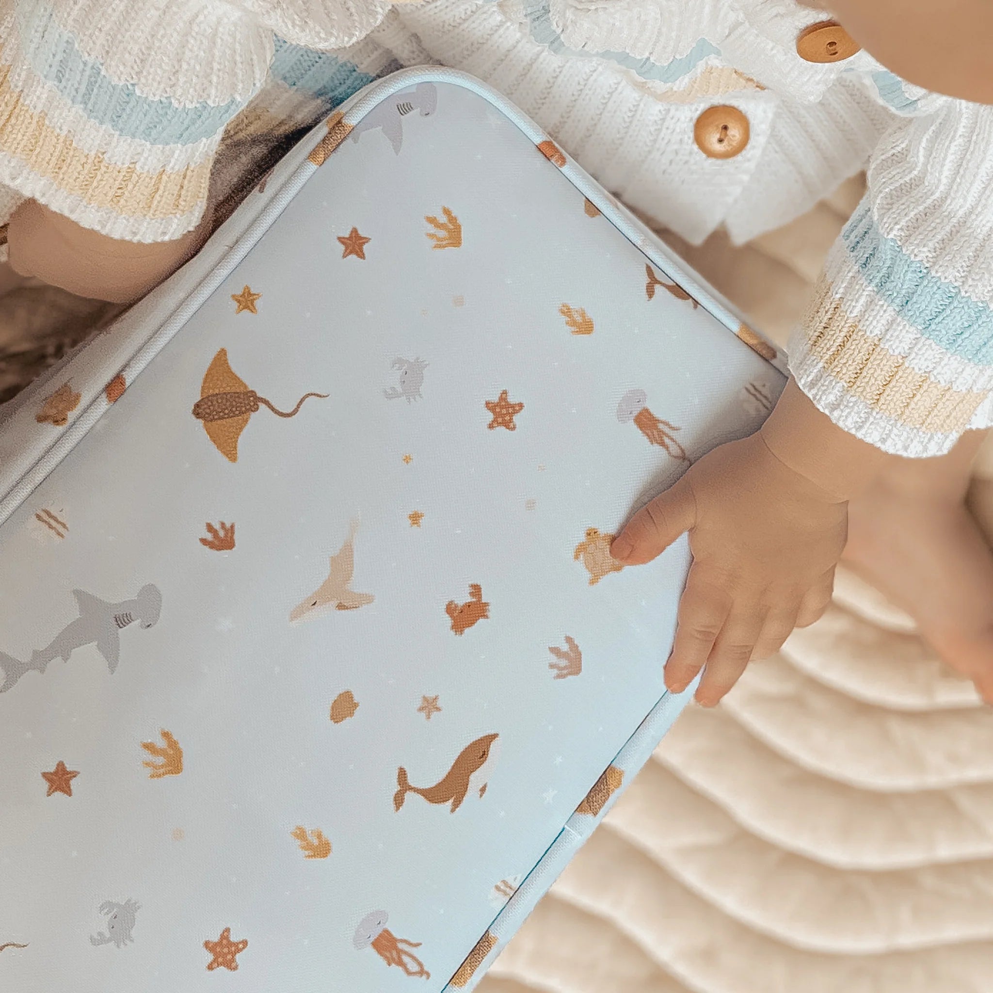 A baby with light skin wearing a white sweater with blue and beige stripes and wooden buttons is sitting on a beige mat. They are holding the Ocean Creatures Sky Lunch Bag by FOX & FALLOW, which is made of waterproof fabric and decorated with sea-themed illustrations like whales, starfish, and jellyfish.