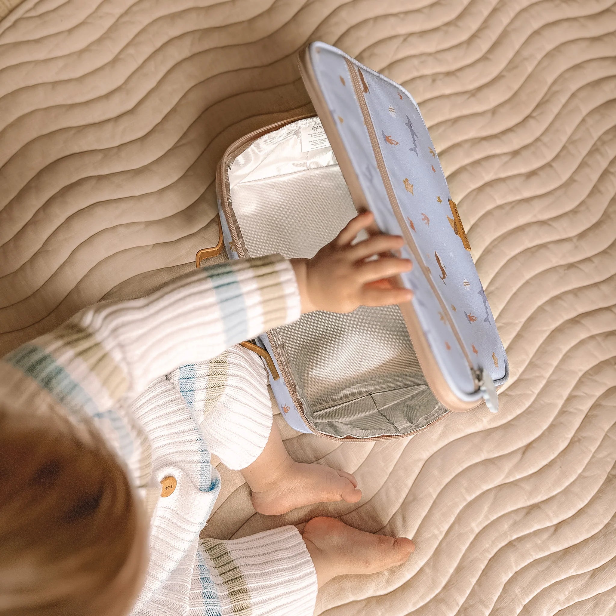 A toddler in striped pajamas sits on a textured beige blanket, reaching into an Ocean Creatures Sky Lunch Bag by FOX & FALLOW with a pale blue outer surface and reflective silver interior. The kid-approved lunch bag lid features cute animal prints, including small birds and stars.