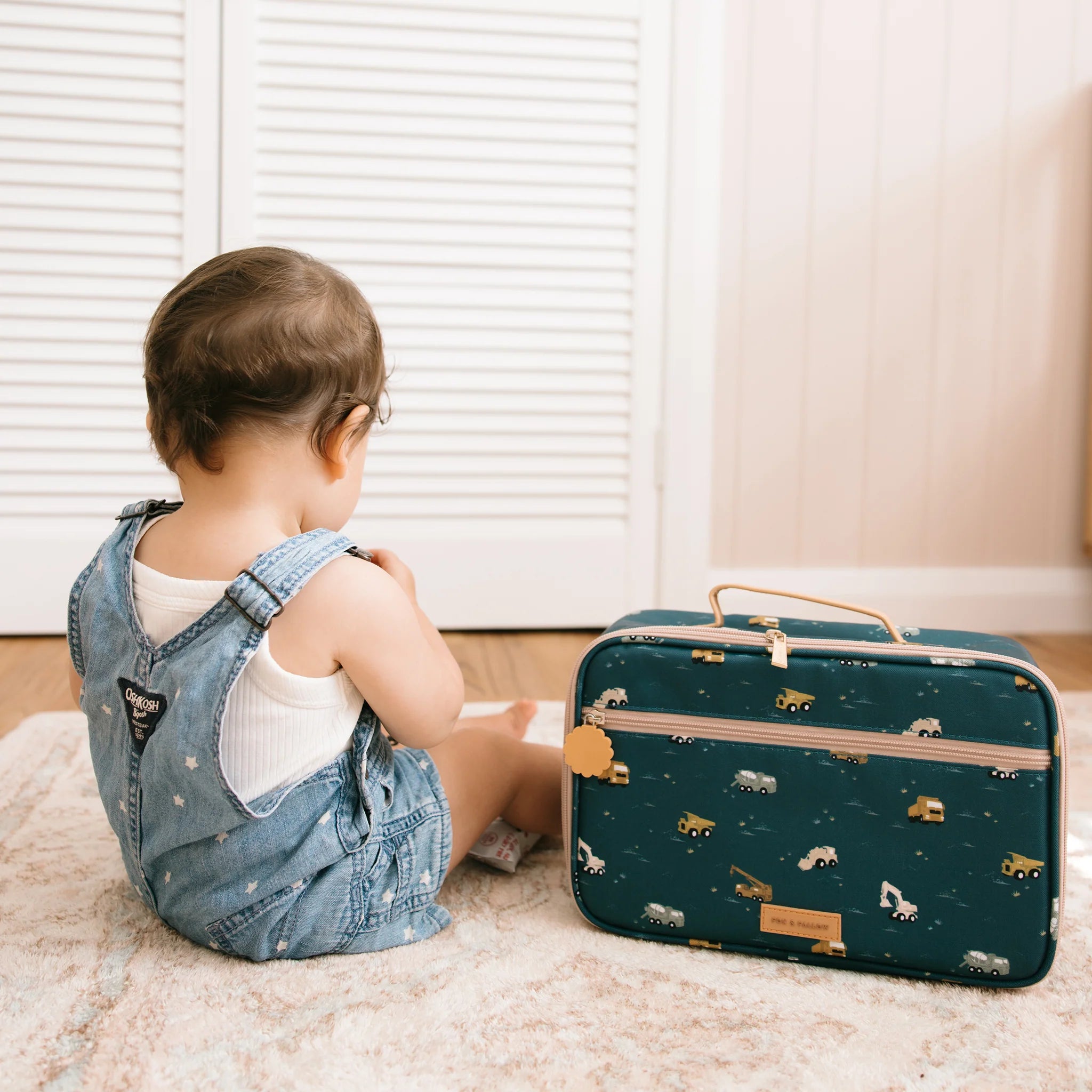 A toddler in denim overalls sits on a rug, facing the opposite direction. Beside them is a small, dark-colored FOX & FALLOW Trucks Lunch Bag featuring a whimsical vehicle pattern made from easy-to-clean, waterproof fabric. The backdrop includes white louvered doors.