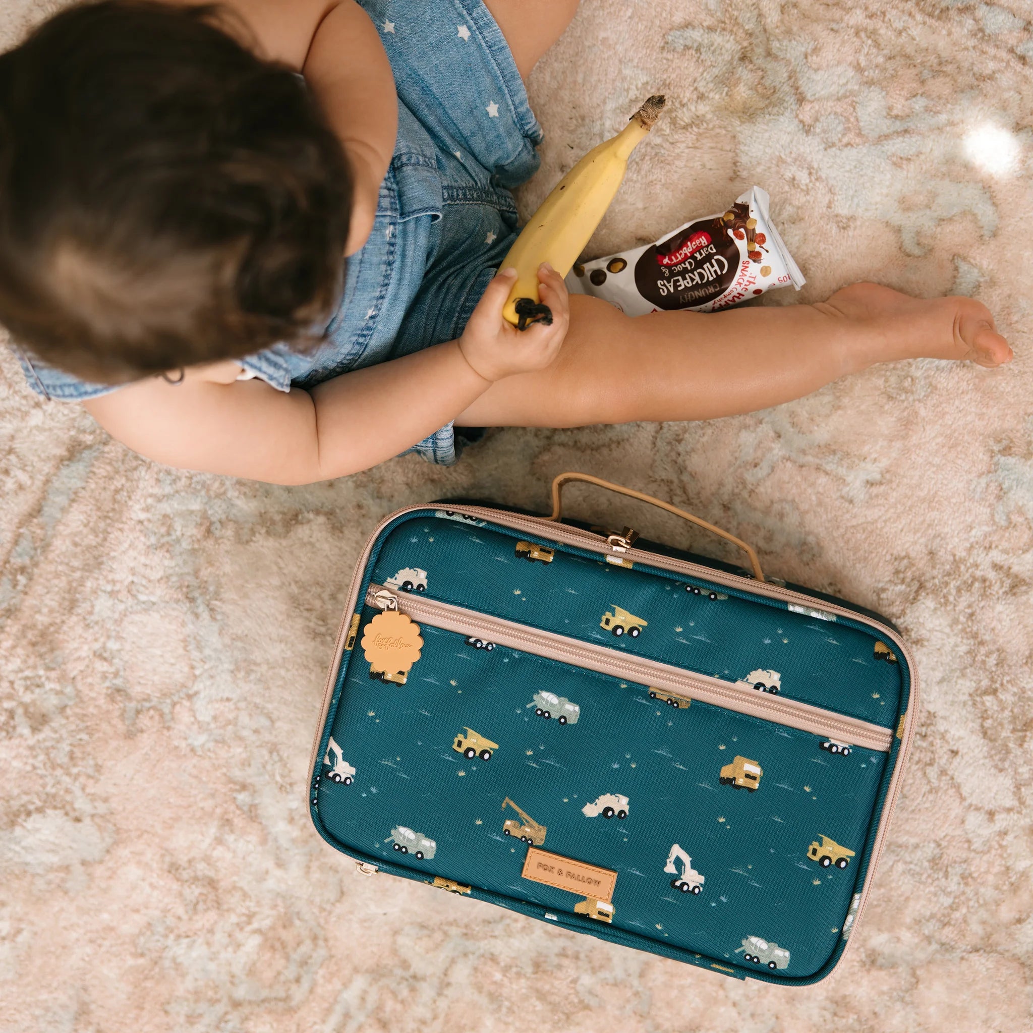 A child sits on a carpet, holding a banana and a snack packet. Beside them is an easy-to-clean Trucks Lunch Bag from FOX & FALLOW, featuring vehicle designs and made from waterproof fabric. They are wearing a denim outfit.