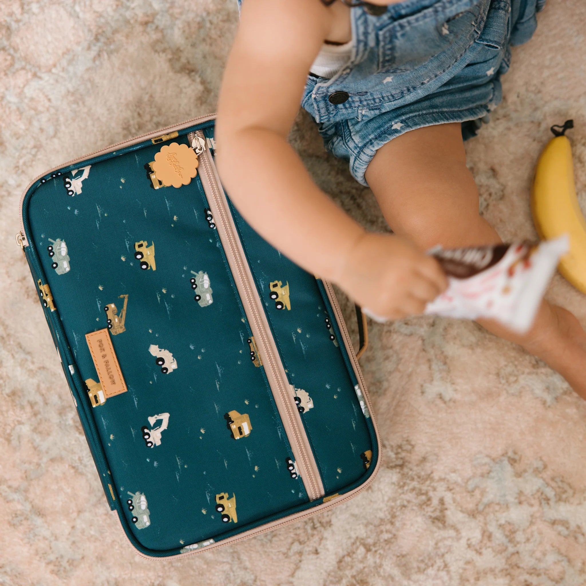 A toddler in denim shorts sits on a patterned carpet, reaching for a snack from the Trucks Lunch Bag by FOX & FALLOW. Next to them is a teal suitcase featuring cute animal and vehicle designs. The waterproof fabric makes it easy to clean, and there's a banana lying nearby.