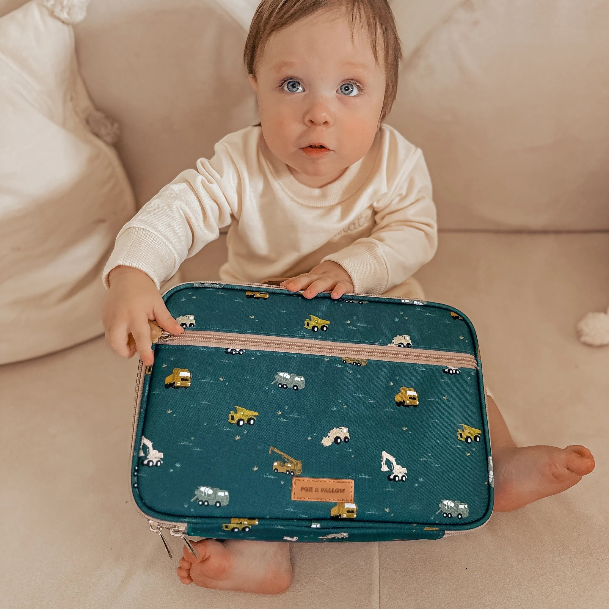 A baby sits on a beige couch, holding a blue-green FOX & FALLOW Trucks Lunch Bag crafted from waterproof fabric. The lunch bag, adorned with construction vehicles and a label with text, is easy to clean. The baby, dressed in a cream outfit, gazes upward with a curious expression.