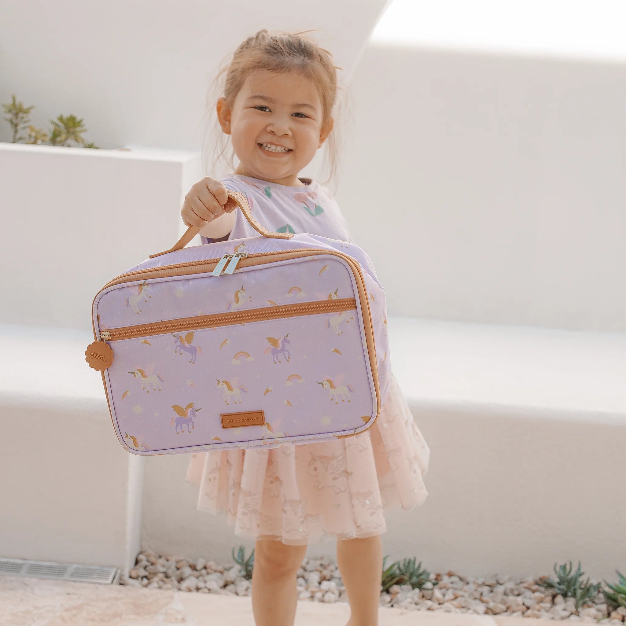 A smiling young girl holds up the FOX & FALLOW Unicorns Lunch Bag, which is popular with kids for its insulation. She is wearing a pink dress and standing outdoors on a stone surface with succulents in the background.