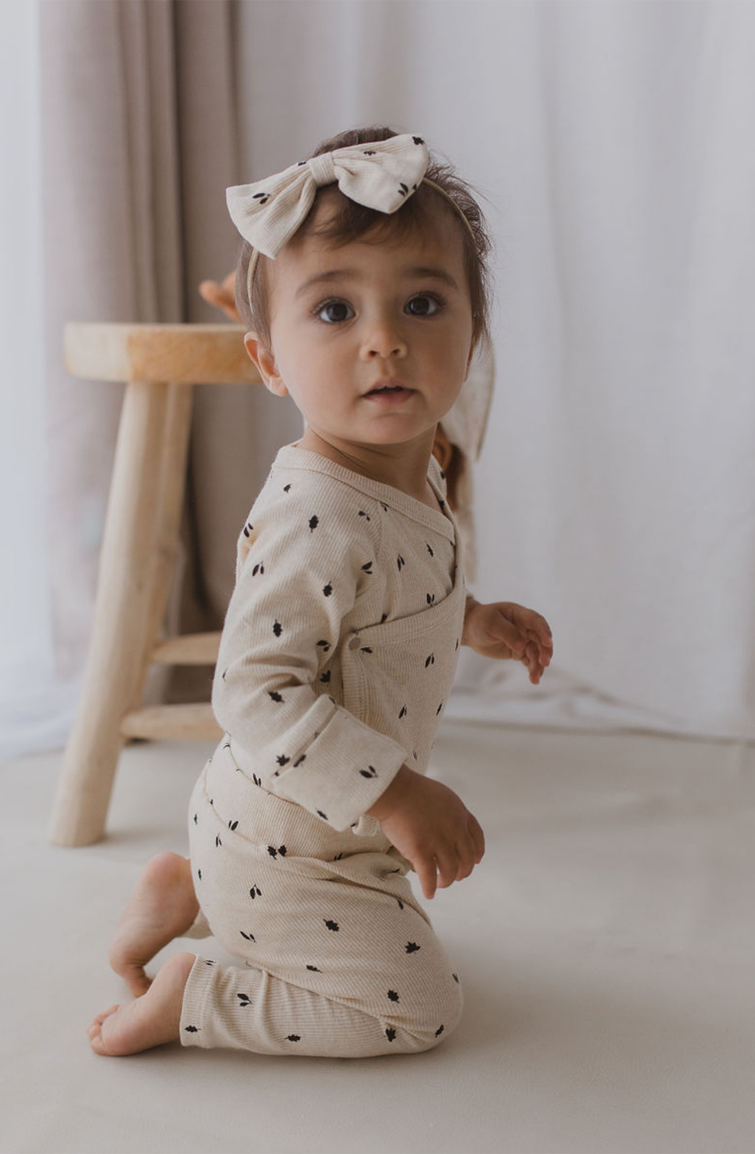 A small child with short hair, wearing a pair of SUSUKOSHI Drawstring Leggings Fall Leaves and a matching headband with a bow, is kneeling on the floor. In the background, part of a wooden stool and white curtains are visible. The ethically made outfit from organic cotton ensures both comfort and sustainability.