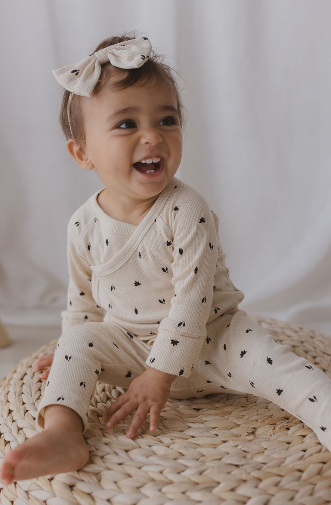 A toddler with short brown hair smiles joyfully while sitting on a woven mat. The child is wearing SUSUKOSHI's ethically made Drawstring Leggings Fall Leaves, a cream-colored outfit adorned with small black prints, and a large cream bow on their head. A white curtain serves as the backdrop.