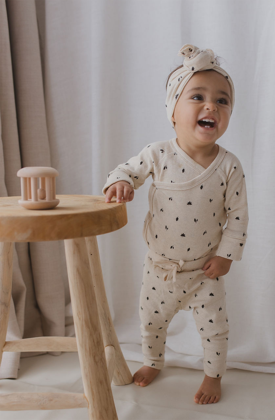 A smiling toddler stands next to a wooden stool, dressed in SUSUKOSHI's matching patterned outfit made from organic cotton called Drawstring Leggings Fall Leaves and a headband. The toddler's light-colored outfit featuring small black prints reflects its ethically made origins. A beige-colored toy rests on the stool, with a light-colored curtain in the background.