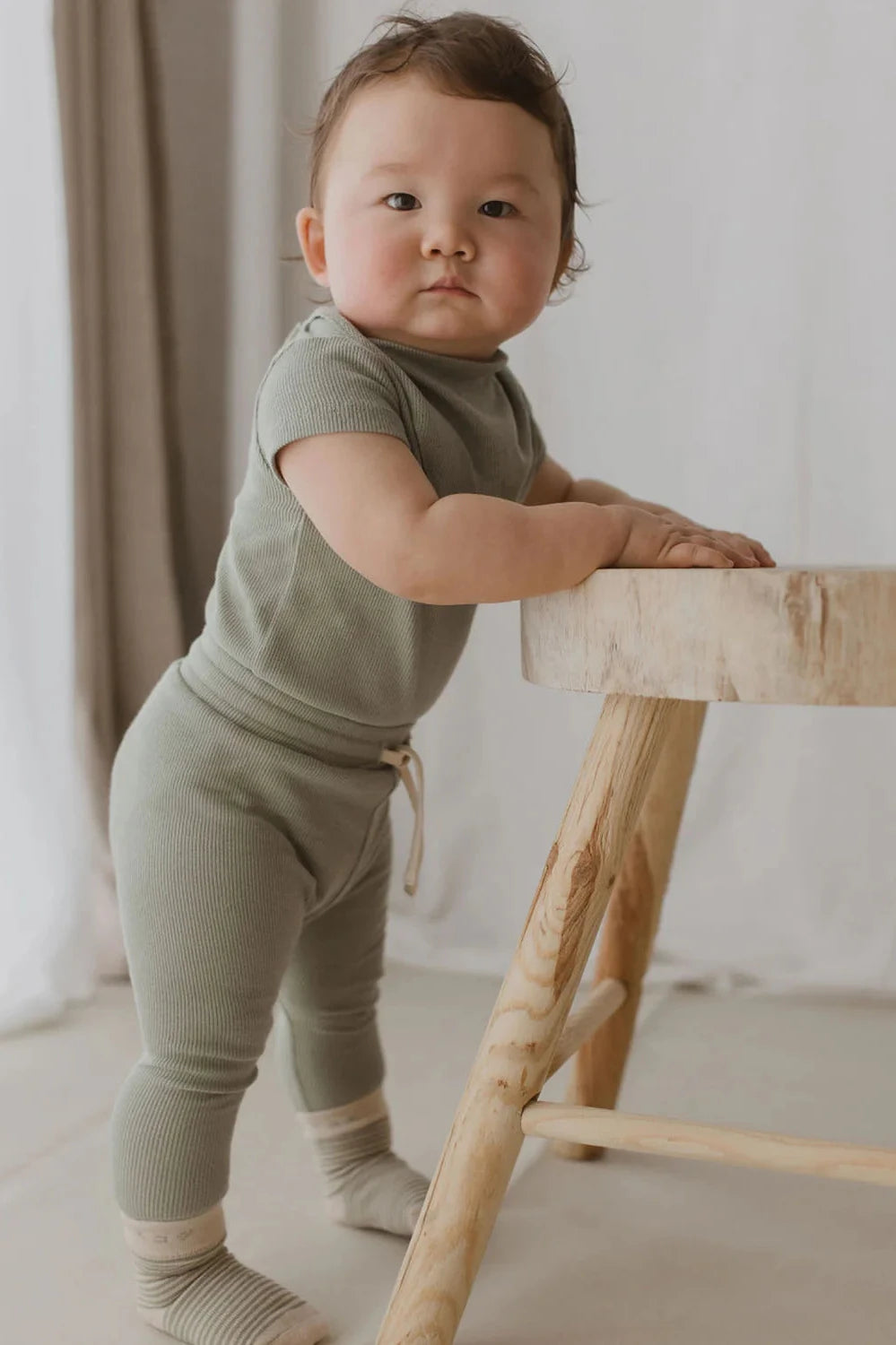 A toddler in SUSUKOSHI's Drawstring Legging Sage (Size 0-3M), made from ethically sourced organic cotton, stands leaning on a round wooden stool. The child, with light brown hair and rosy cheeks, wears matching socks. Soft white and beige curtains adorn the background.