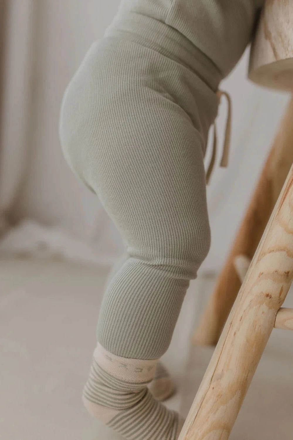 A close-up of a baby standing and holding onto a wooden chair, wearing the "Drawstring Legging Sage" by SUSUKOSHI and beige socks. The background is softly blurred, creating a warm and cozy atmosphere.