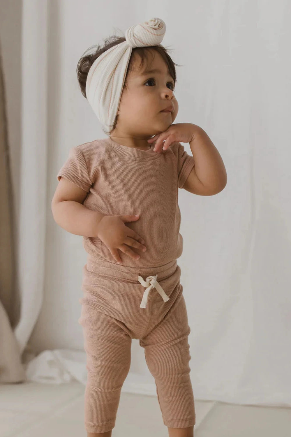 A toddler stands indoors wearing a matching terracotta outfit, including the Drawstring Legging from SUSUKOSHI, which features an ethically made short-sleeved top and leggings. They are adorned with a cream-colored headband with a knot, gazing slightly upward while thoughtfully touching their chin.