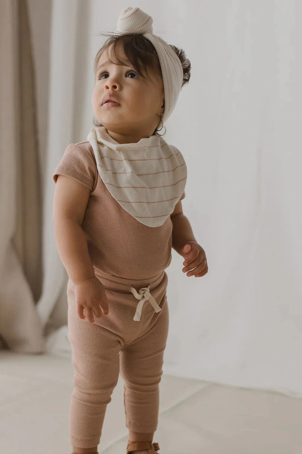 A toddler dressed in the SUSUKOSHI Drawstring Legging Terracotta, complete with a matching headband and striped bandana, gazes upward. The softly blurred background features light-colored curtains, and the child stands on a light-hued floor.