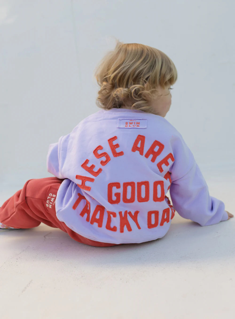 A young child with blond hair, seen from behind, wears the Small Swim Club - Good Tracky Pullover in lavender/red by SMALL SWIM CLUB and matching red fleece-lined Tracky Dacks with "THESE ARE GOOD TRACKY DAYS" text. The background is plain and light-colored.