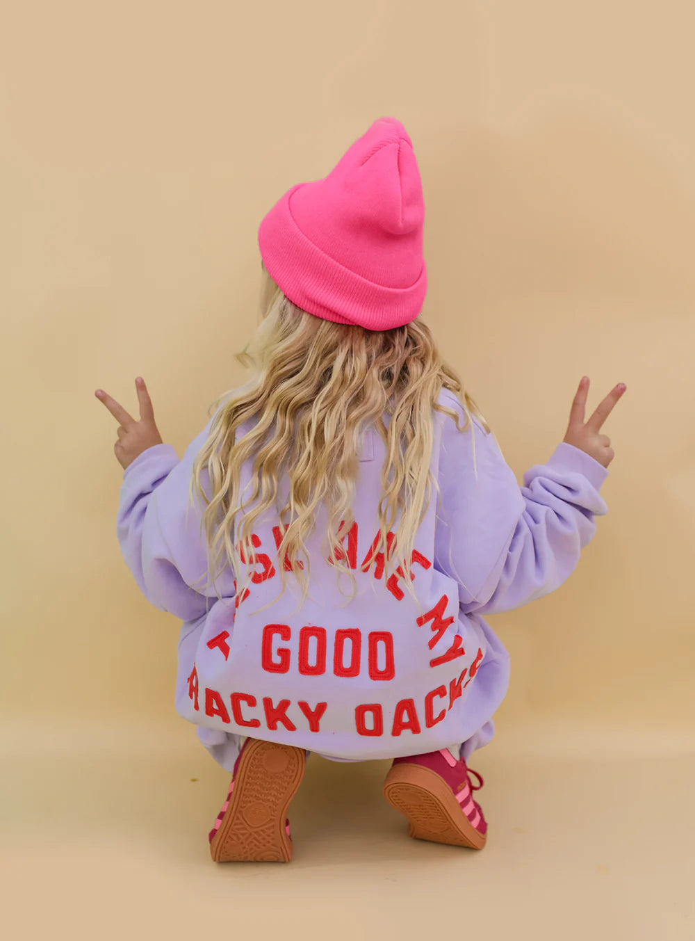 A child in a bright pink beanie and SMALL SWIM CLUB Good Tracky Pullover Lavender/Red squats facing away, making peace signs with both hands against a beige background.