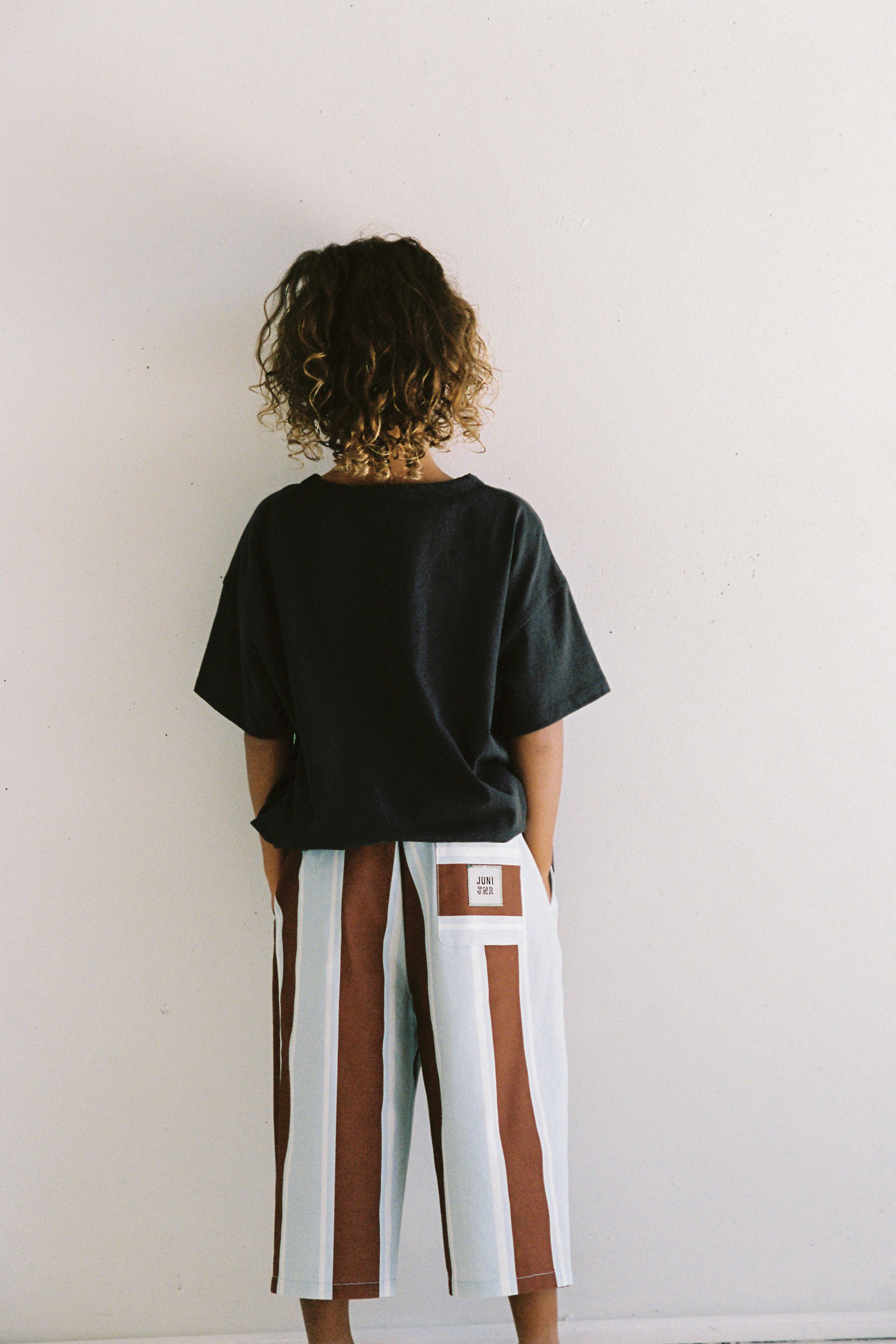 A child with curly hair stands facing away from the camera against a plain white wall. They are wearing a black T-shirt and knee-length Lenny Pant Watson Stripe shorts by JUNI JUNIOR, featuring vertically striped patterns in shades of blue and brown with an elastic waistband for comfort. The child has their hands in the pockets of their shorts.