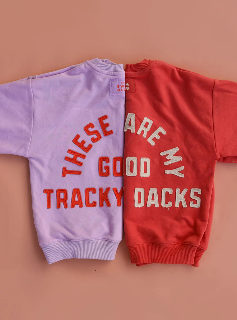Two SMALL SWIM CLUB children's sweatshirts, including the Small Swim Club - Good Tracky Pullover in Red/Cream, are side by side on a pink surface; their varsity style text forms a playful phrase when paired.