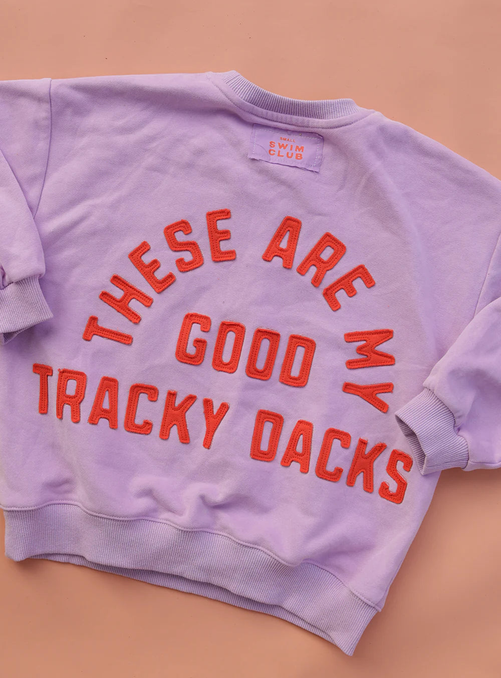 The SMALL SWIM CLUB Small Swim Club - Good Tracky Pullover Lavender/Red, a slouchy-fit lavender sweatshirt with red and orange "THESE ARE MY GOOD TRACKY DACKS" text on the back, is shown laid flat on a peach-colored surface.