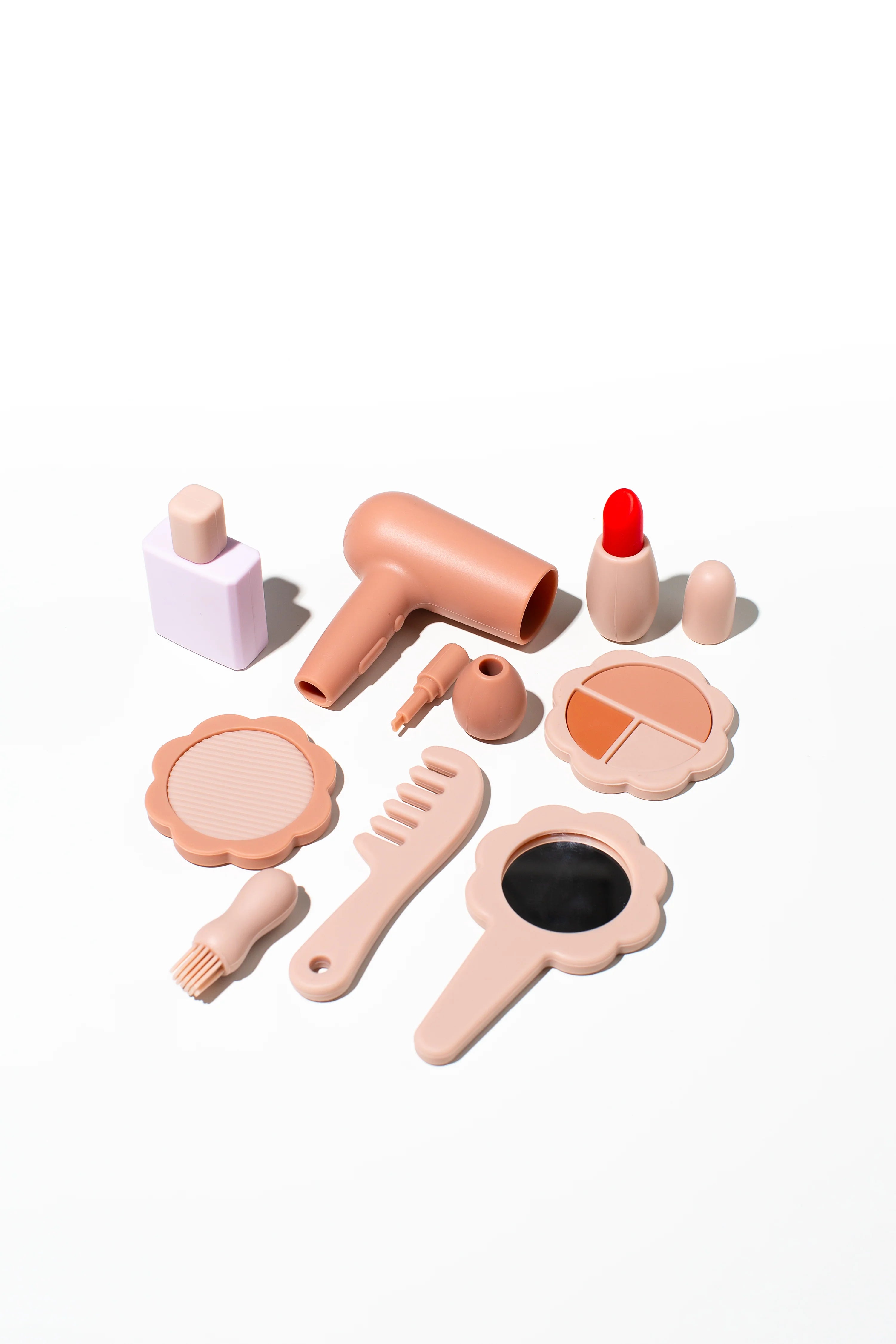 The LITTLE DROP Beauty Set features an adorable kids' beauty kit arranged on a white surface, including a toy hairdryer, lipstick, mirror, perfume bottle, makeup palette, comb, brush, and other small beauty tools in shades of beige and light pink. These silicone beauty toys inspire creativity and fun.