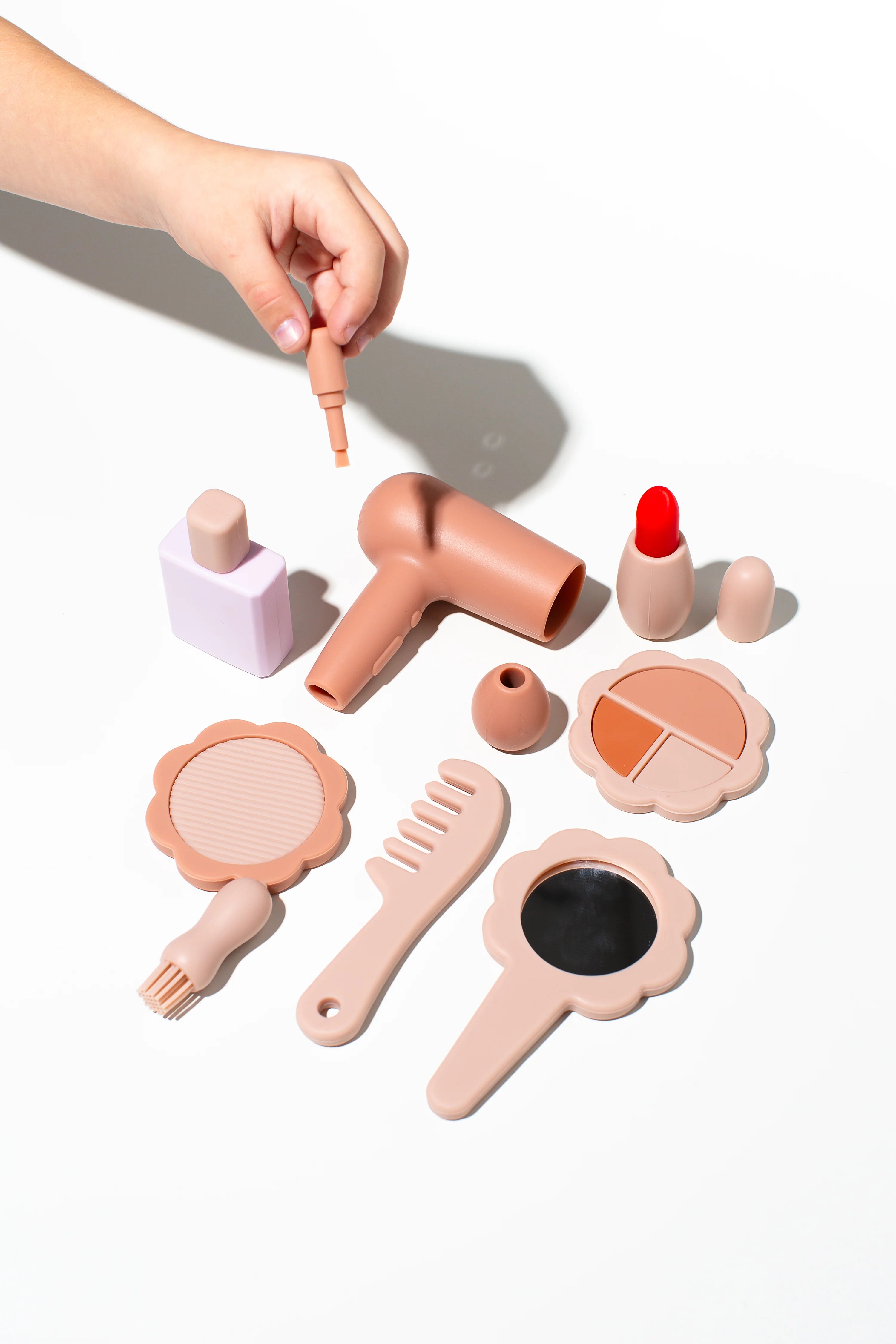 A child's hand reaches for a toy makeup brush from an arrangement of LITTLE DROP Beauty Set items, including a hair dryer, lipstick, blush compact, perfume bottle, comb, mirror, and other silicone beauty toys in pastel shades laid out on a white surface.