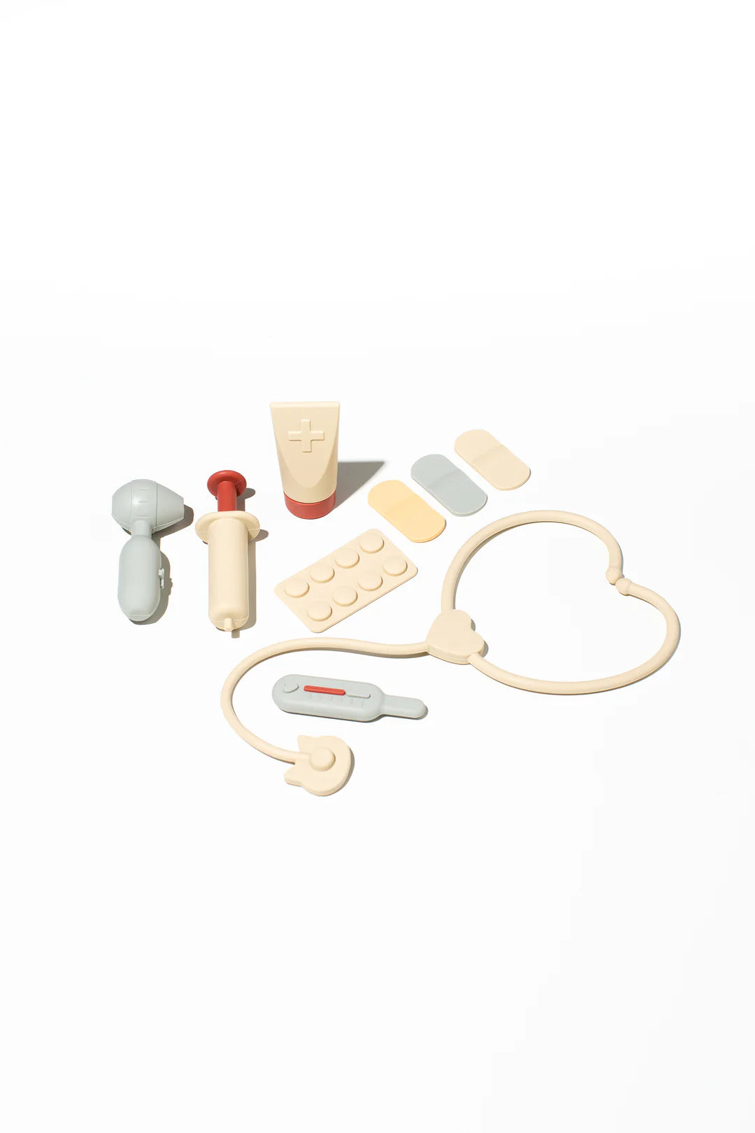 The LITTLE DROP Doctor Set on a white background features a stethoscope, thermometer, syringe, medicine bottle, tube of ointment, two adhesive bandages, and a collection of tablets. Predominantly beige with red and gray accents, this silicone playset is perfect for sparking a child's imagination.