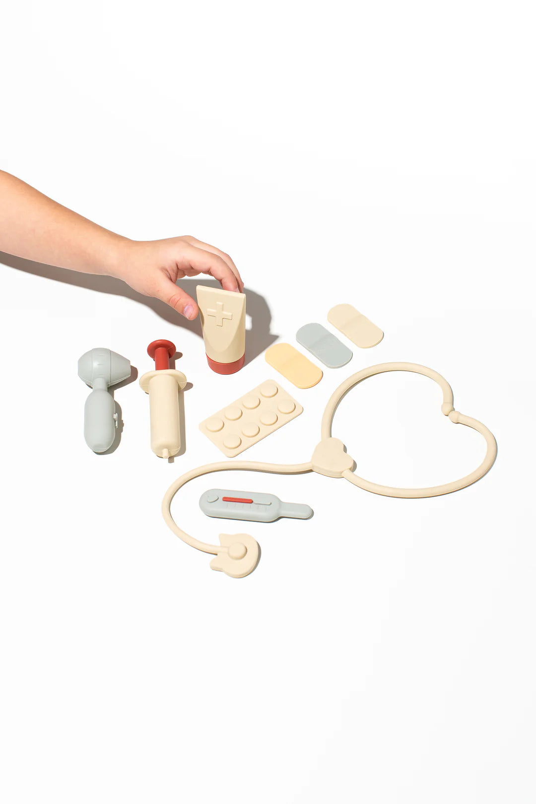 A child's hand reaches for a toy medical cream tube from the LITTLE DROP Doctor Set, nestled amongst a silicone playset that includes a stethoscope, syringe, pill bottle, bandages, and thermometer. This collection nurtures a child's imagination as they delve into their interest in healthcare against a white background.