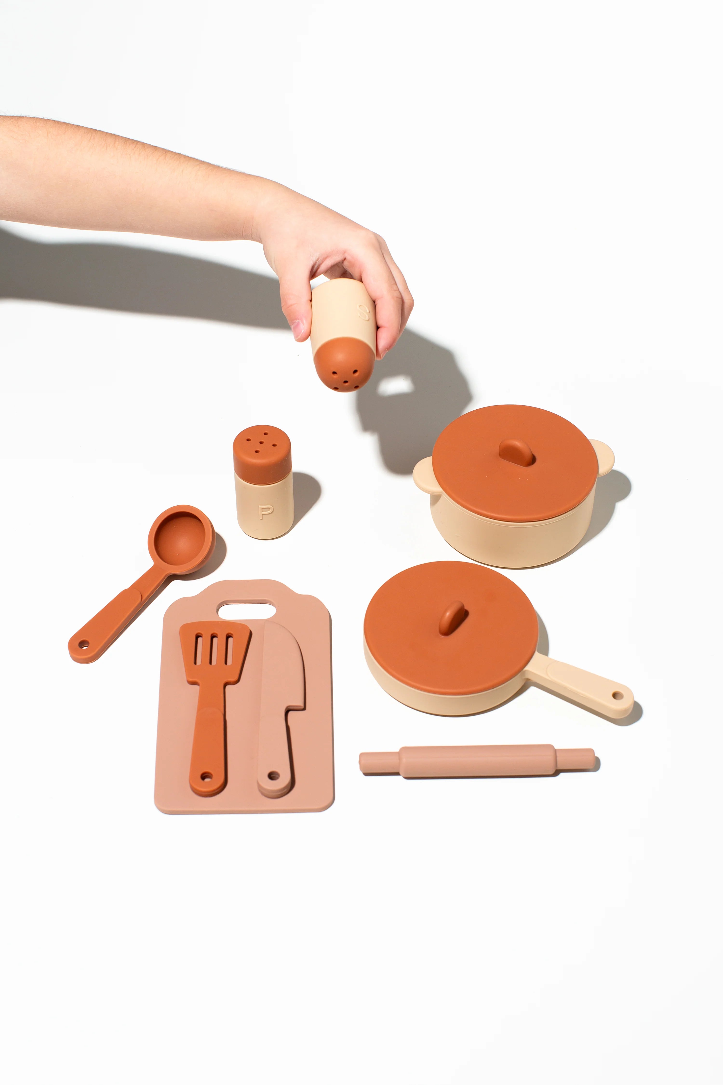 A child's hand holds a felt-tipped shaker above a LITTLE DROP Cooking Set, showcasing kids' culinary skills. The set, in muted earthy tones, includes two saucepans with lids and durable silicone utensils like a rolling pin and cutting board, all neatly arranged on a white background.