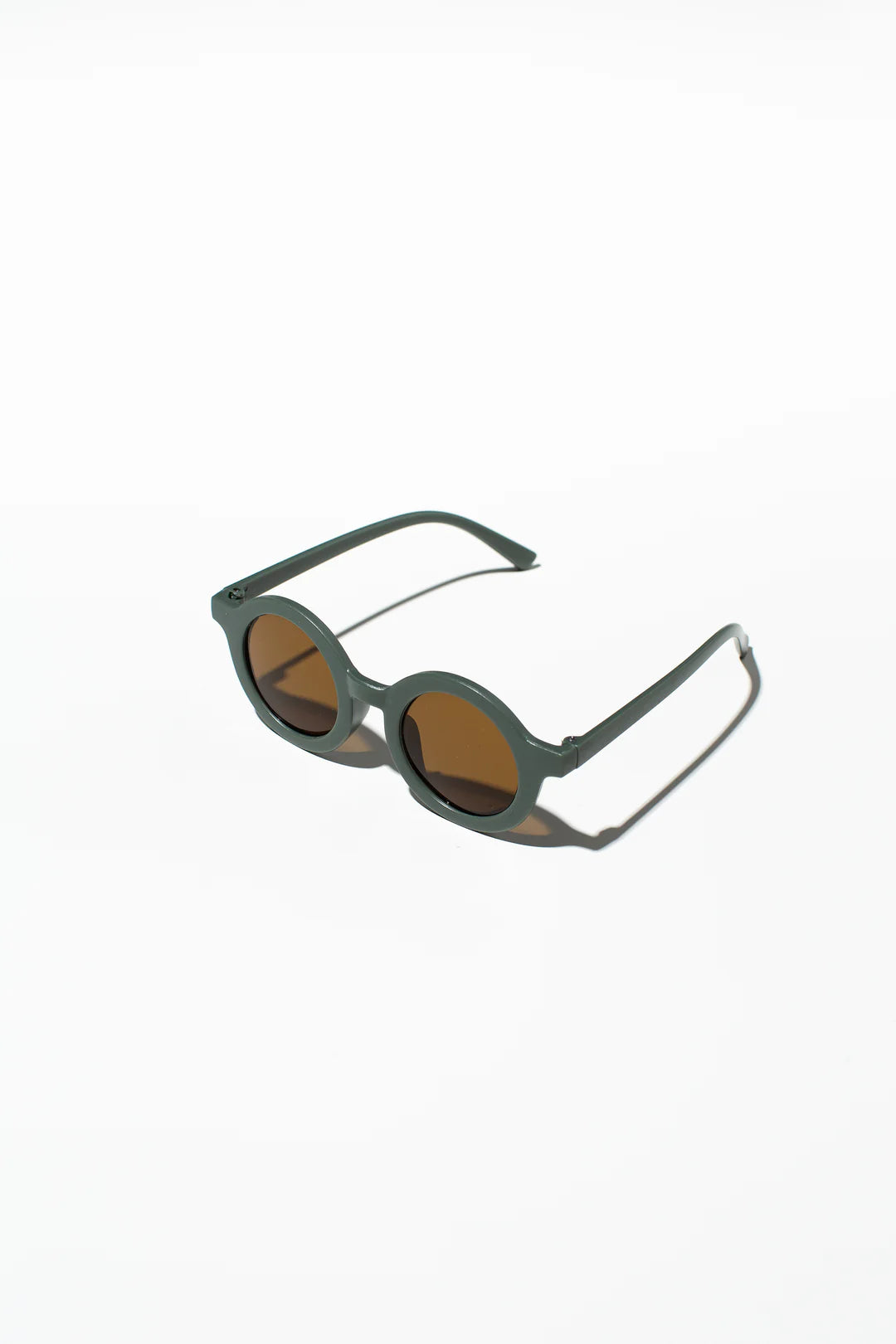 A pair of LITTLE DROP Kids Shades Sage with dark green frames and brown lenses is placed on a white surface. The temples are extended, casting a faint shadow. The design is minimalist and modern, and they come with UV400 protection for optimal eye safety.