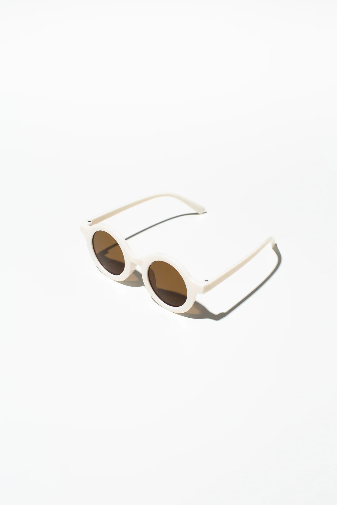 A pair of Kids Shades Bone from LITTLE DROP, featuring round, white frames and dark lenses, is showcased in a minimalist setting against a white background. The sunglasses, offering UV400 protection, cast a slight shadow to the right.