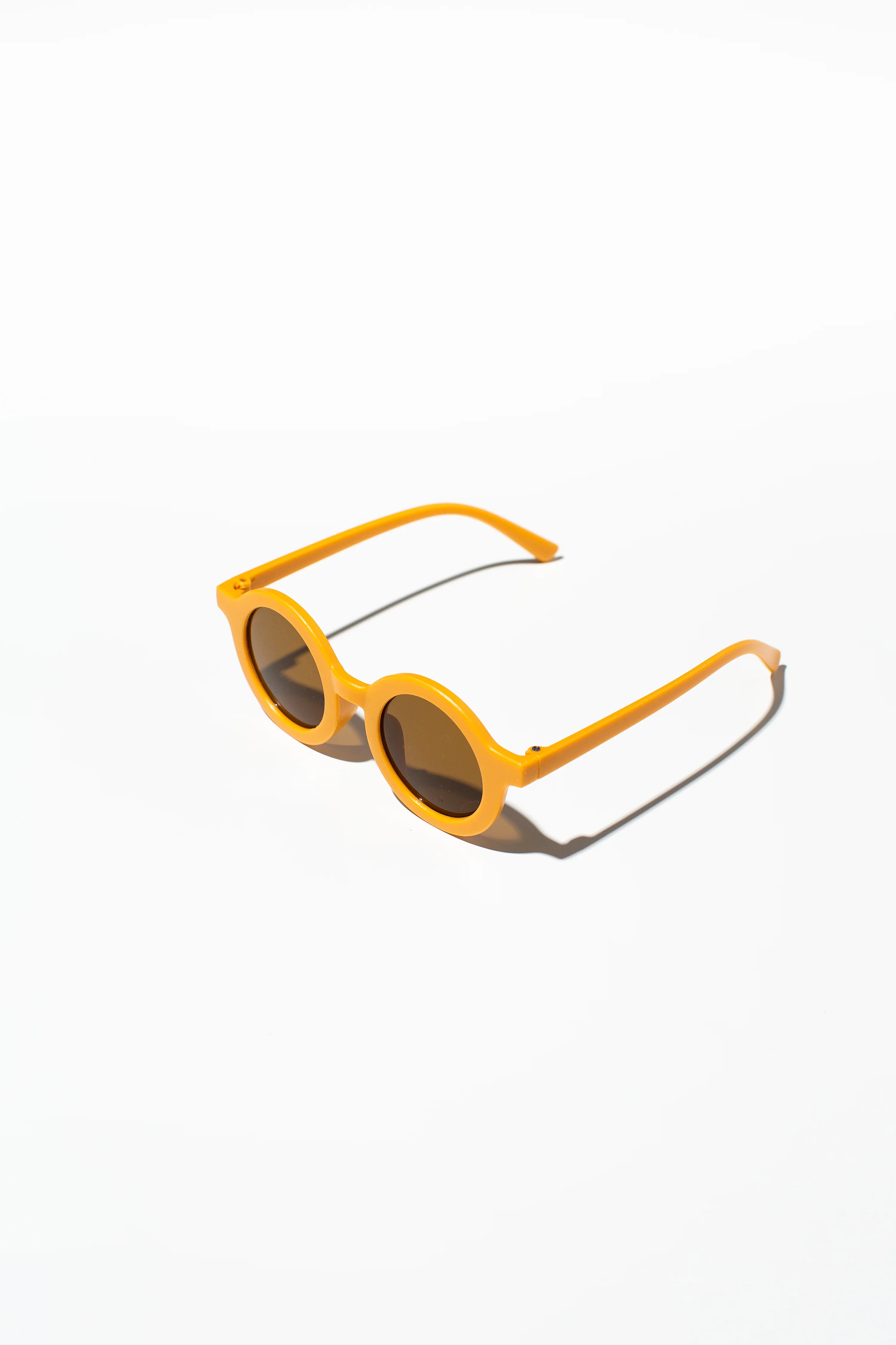 A pair of LITTLE DROP Kids Shades Sand with UV400 protection and dark lenses lies on a white surface, casting subtle shadows to the right.