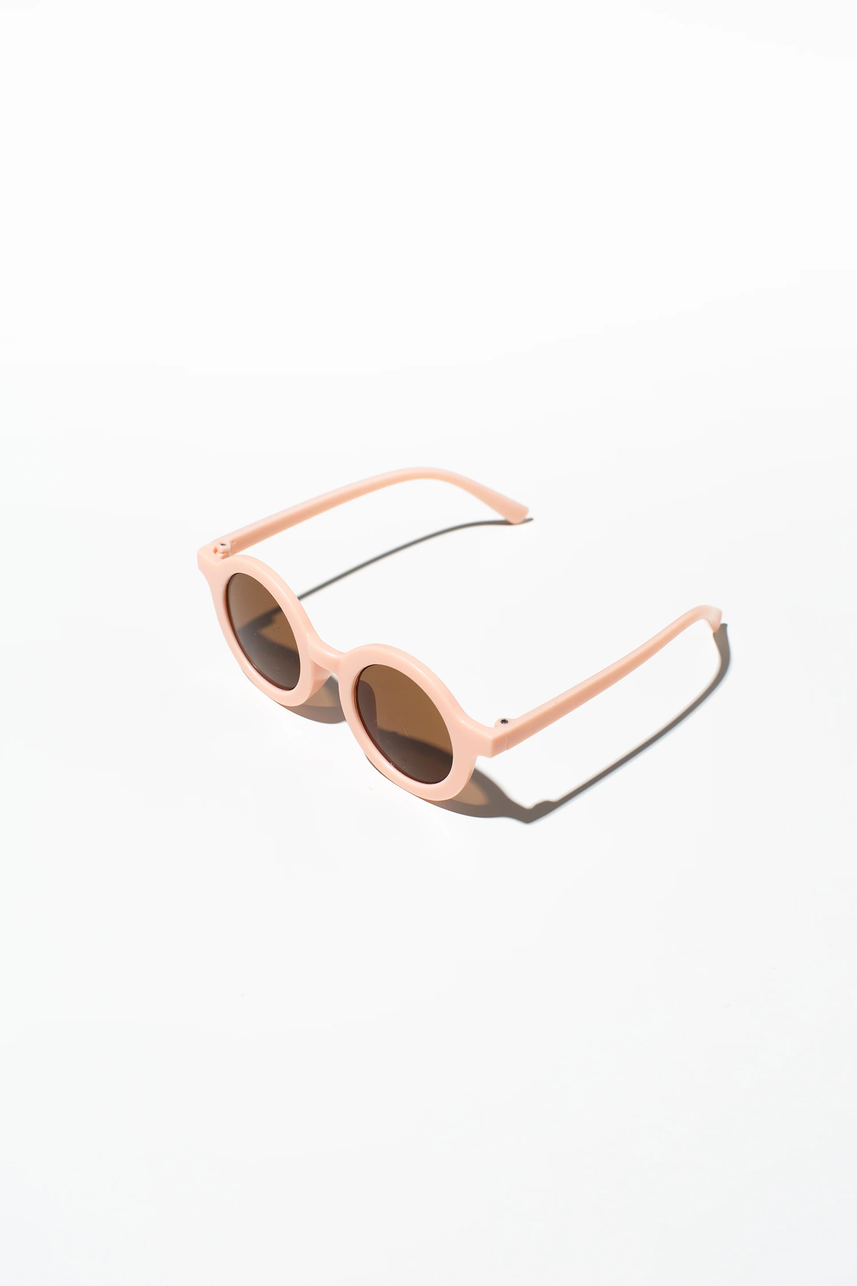 A pair of Kids Shades Peach sunglasses from LITTLE DROP, featuring modern rounded frames in a pale pink hue and dark lenses with UV400 Protection, is placed on a white surface. The sunlight casts faint shadows, enhancing the minimalist and clean aesthetic of the image. Nearby, a silicone sunglasses case completes the stylish ensemble.