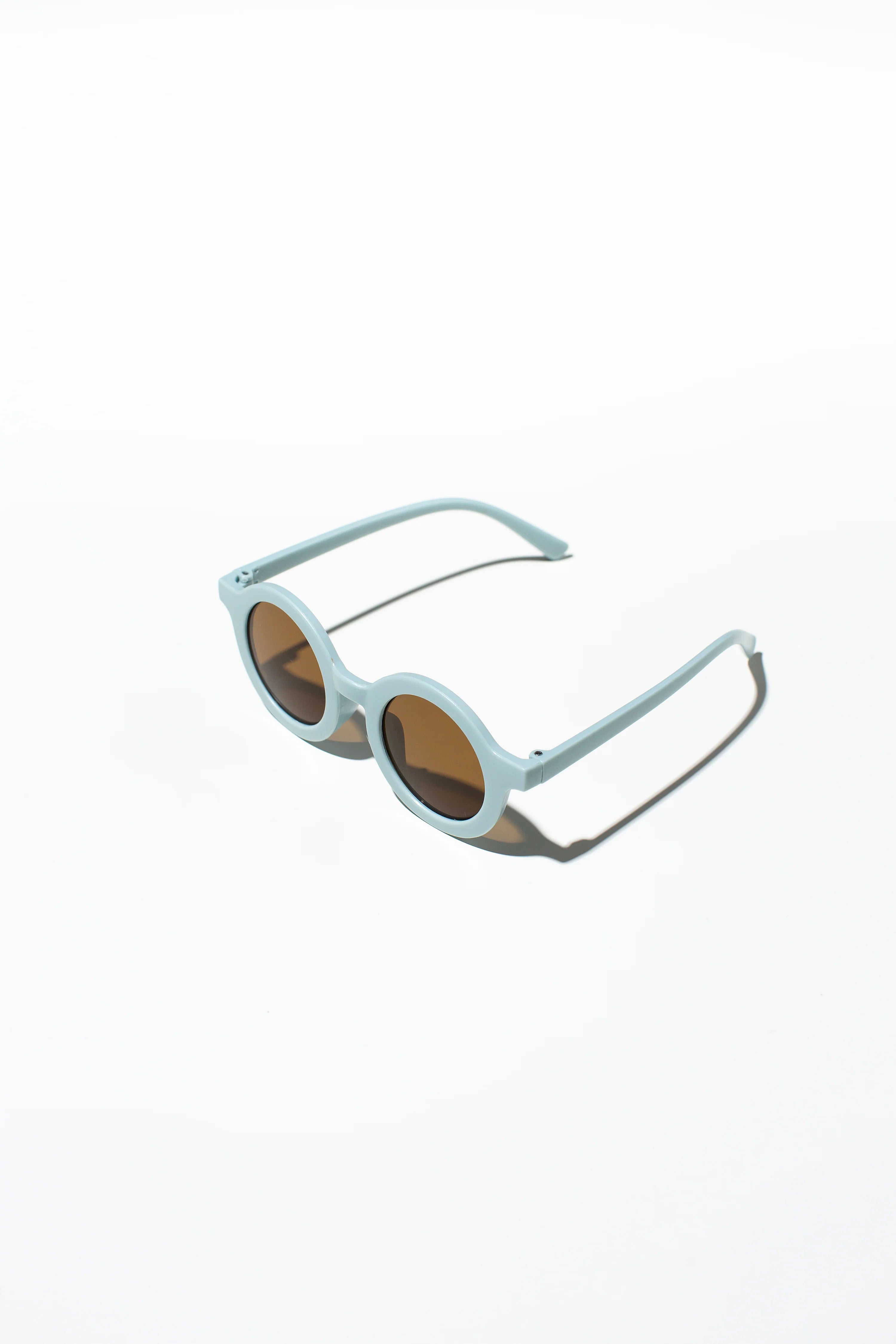 A pair of LITTLE DROP Kids Shades Sky, featuring light blue frames and dark lenses, is placed on a white surface. These round sunglasses have a minimalist design and UV400 protection, with a subtle shadow cast to the right.