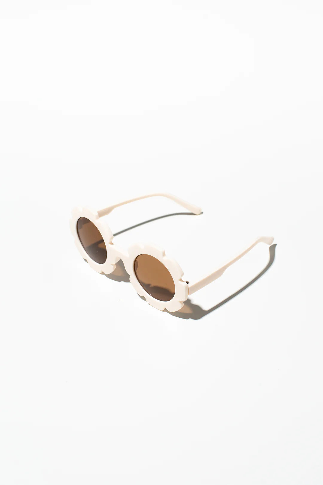 A pair of stylish Daisy Shades Bone kids sunglasses from LITTLE DROP, featuring round lenses and flower-shaped frames, casting soft shadows on a white surface. The dark brown tinted lenses offer UV400 protection.