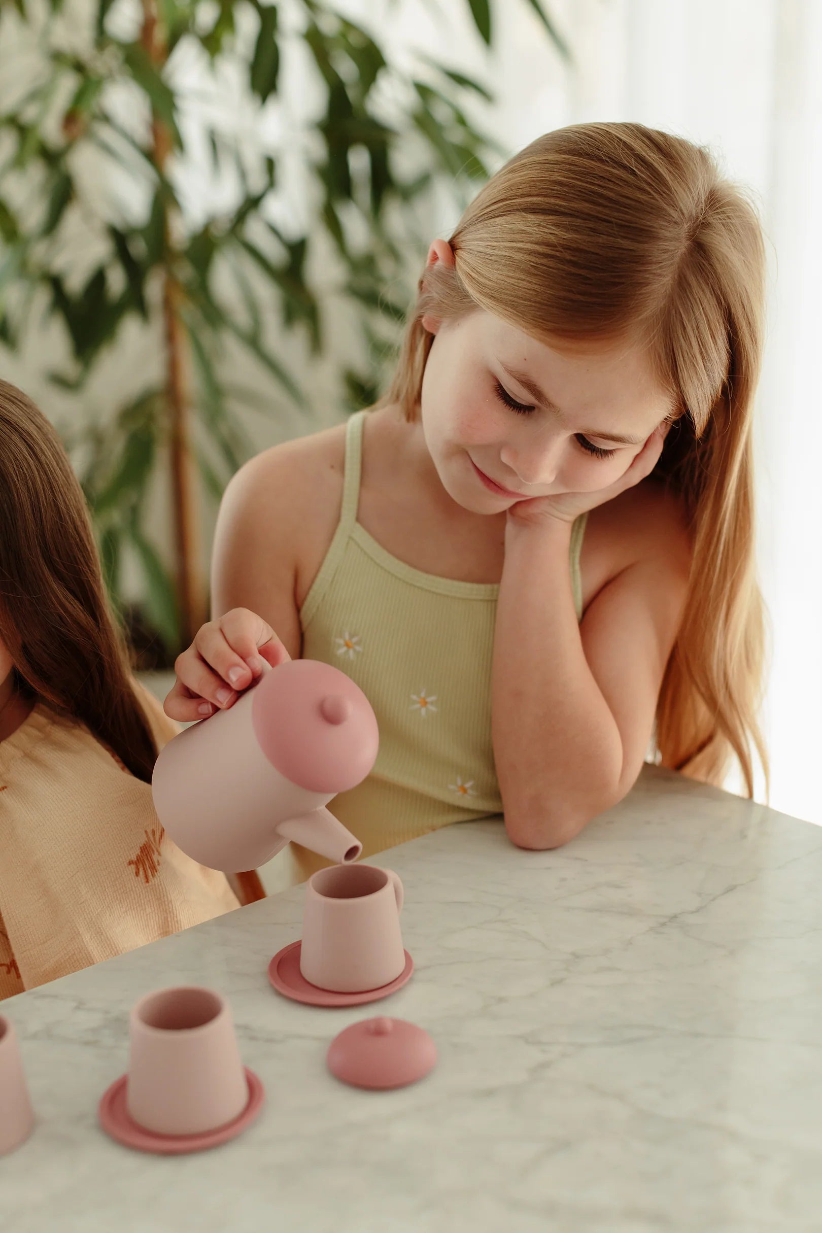 A young girl with long blonde hair, wearing a light green tank top, pours from a small pink teapot into matching cups on the LITTLE DROP Tea Time Set Pink. A green plant is in the background. Another child, partially out of frame, sits beside her.