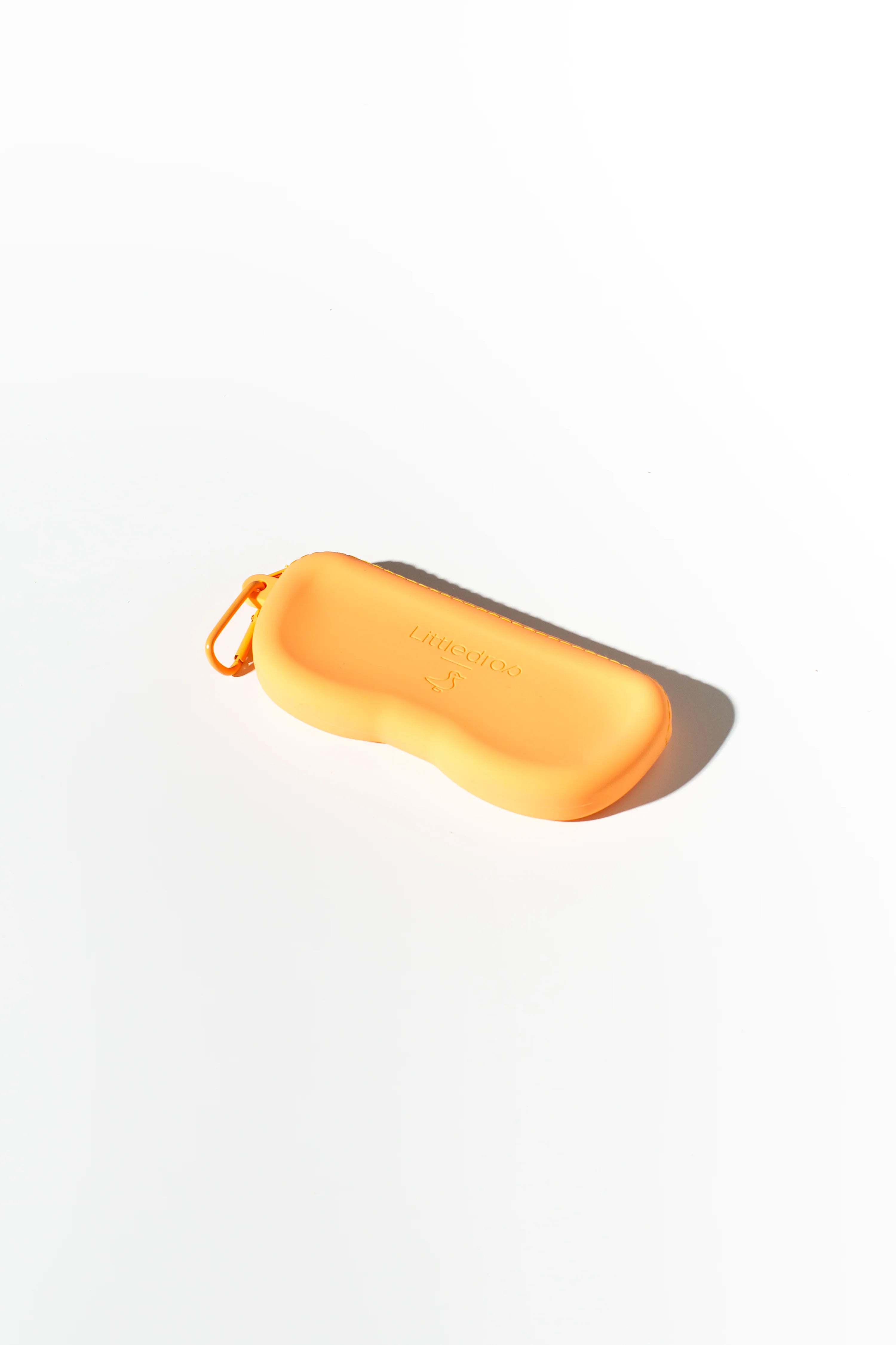 A Kids Shades Bone by LITTLE DROP, an orange, rectangular sunglasses case with rounded edges, lies on a white surface. Designed for children's eyewear, the silicone case features a small carabiner clip on one side and has imprinted text or branding that is not clearly visible.