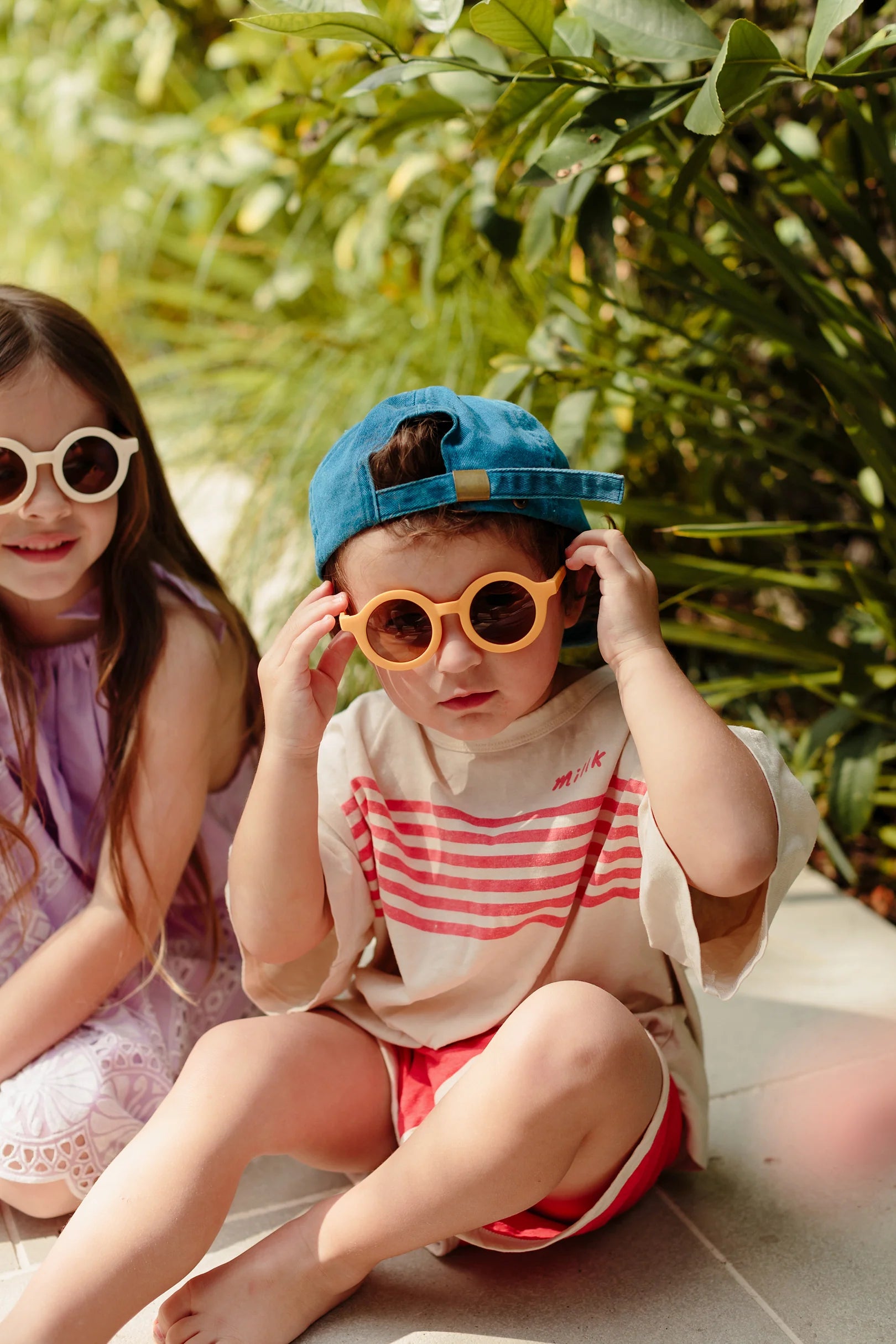 Two children are sitting outdoors in front of greenery. The girl on the left is wearing round white Kids Shades Sand by LITTLE DROP and a purple dress, while the boy on the right is adjusting his orange sunglasses with a blue cap worn backwards. Both have relaxed expressions.