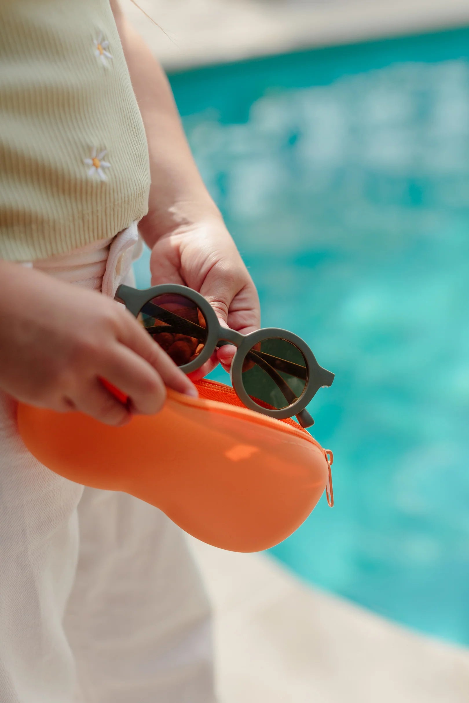 A person stands by a pool, holding an orange half-opened premium silicone sunglasses case and a pair of round, dark Kids Shades Sage by LITTLE DROP. The person is wearing a light-colored top and white pants. The pool water is blue and clear in the background, offering a perfect setting to showcase the Kids Shades Sage's UV400 protection.