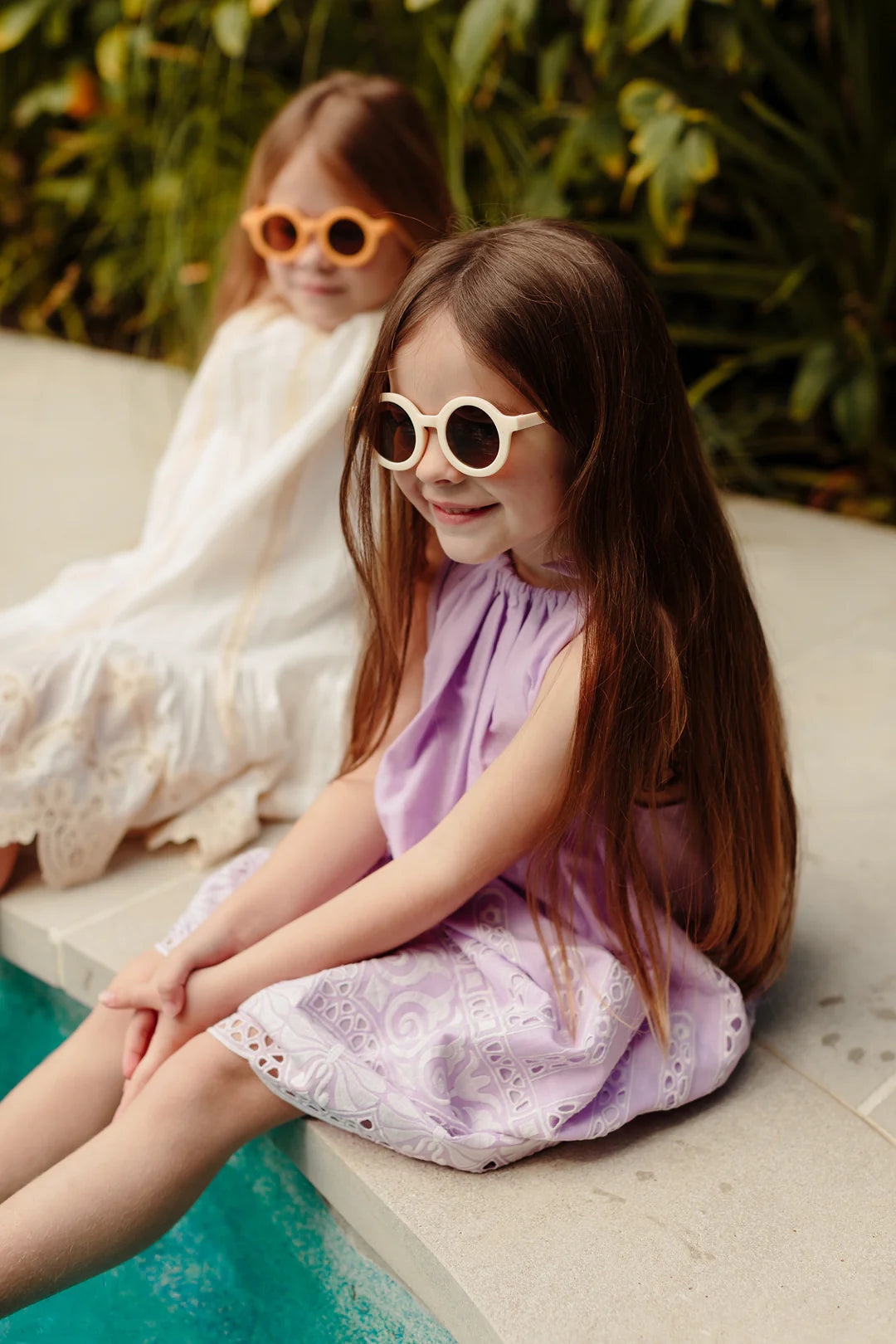 Two young girls sit by the edge of a pool, both donning LITTLE DROP Kids Shades Bone sunglasses with UV400 protection. They have long brown hair; one girl wears a purple dress and the other a white dress. Relaxed and happy, they enjoy a sunny day outdoors with lush greenery in the background.