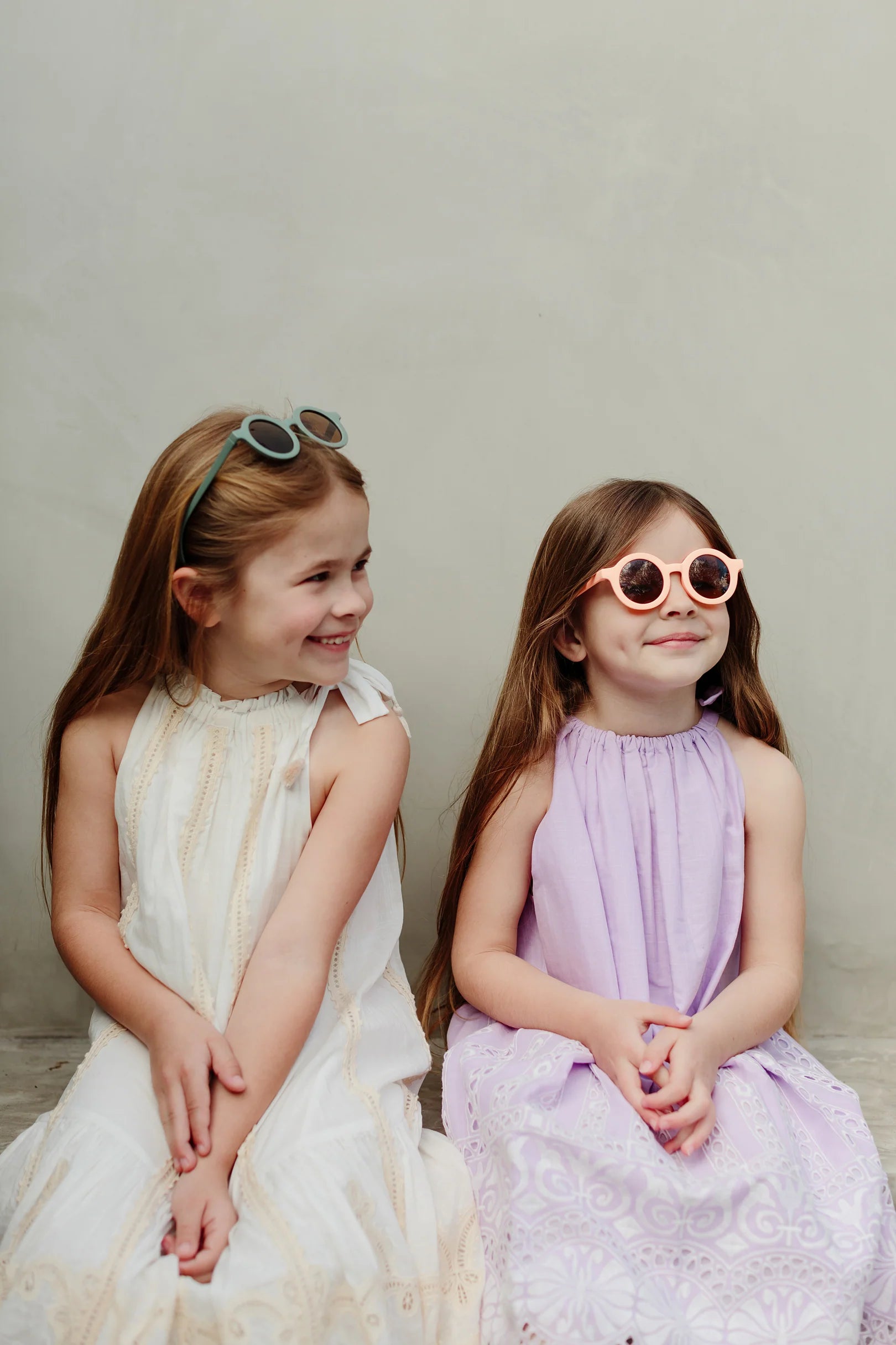 Two young girls with long hair sit together against a plain background. Both are wearing summer dresses, with the girl on the left in a white dress and the girl on the right in a light purple dress and stylish LITTLE DROP Kids Shades Peach offering UV400 protection. They are smiling and looking in opposite directions.