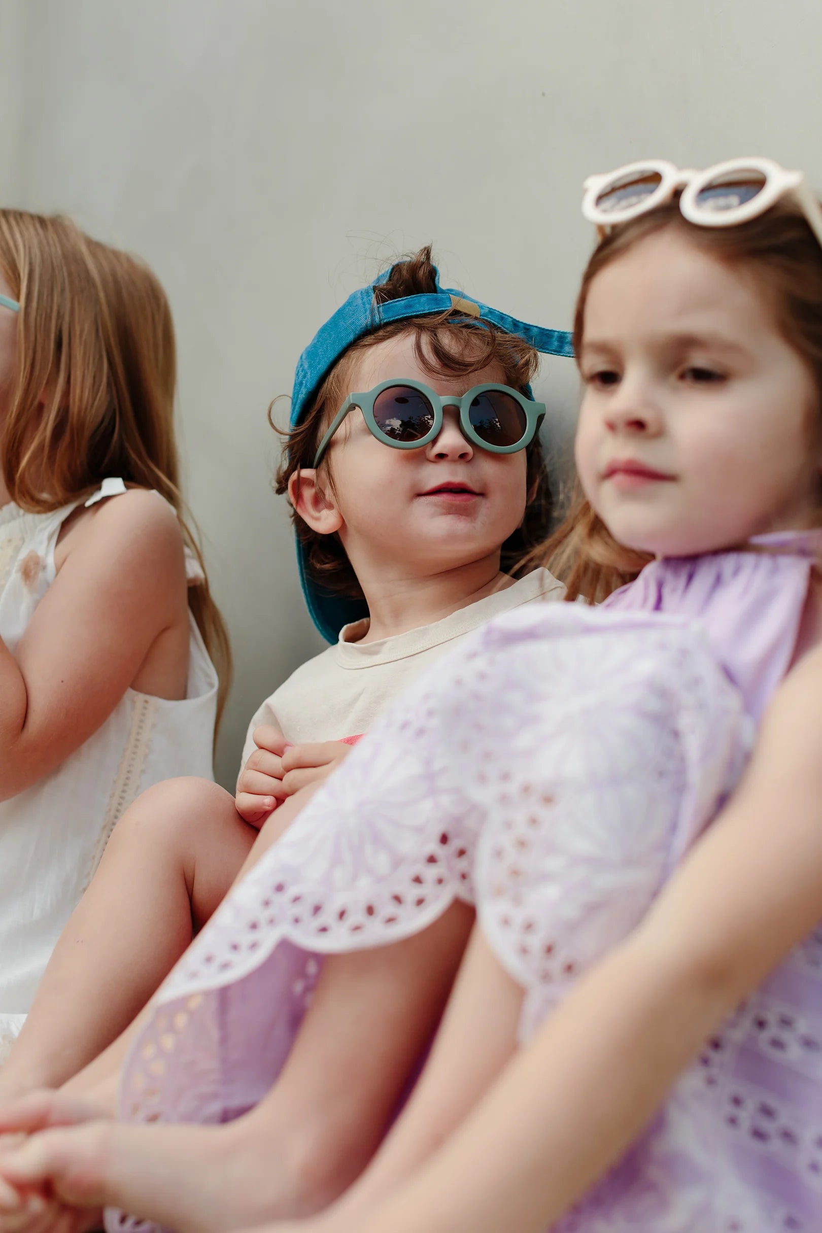 Three young children sit closely together, wearing summer clothes and Kids Shades Sage by LITTLE DROP. The child in the middle sports a blue cap and round sunglasses, while the other two wear white-framed glasses with UV400 Protection. They seem to be outdoors, enjoying a sunny day.