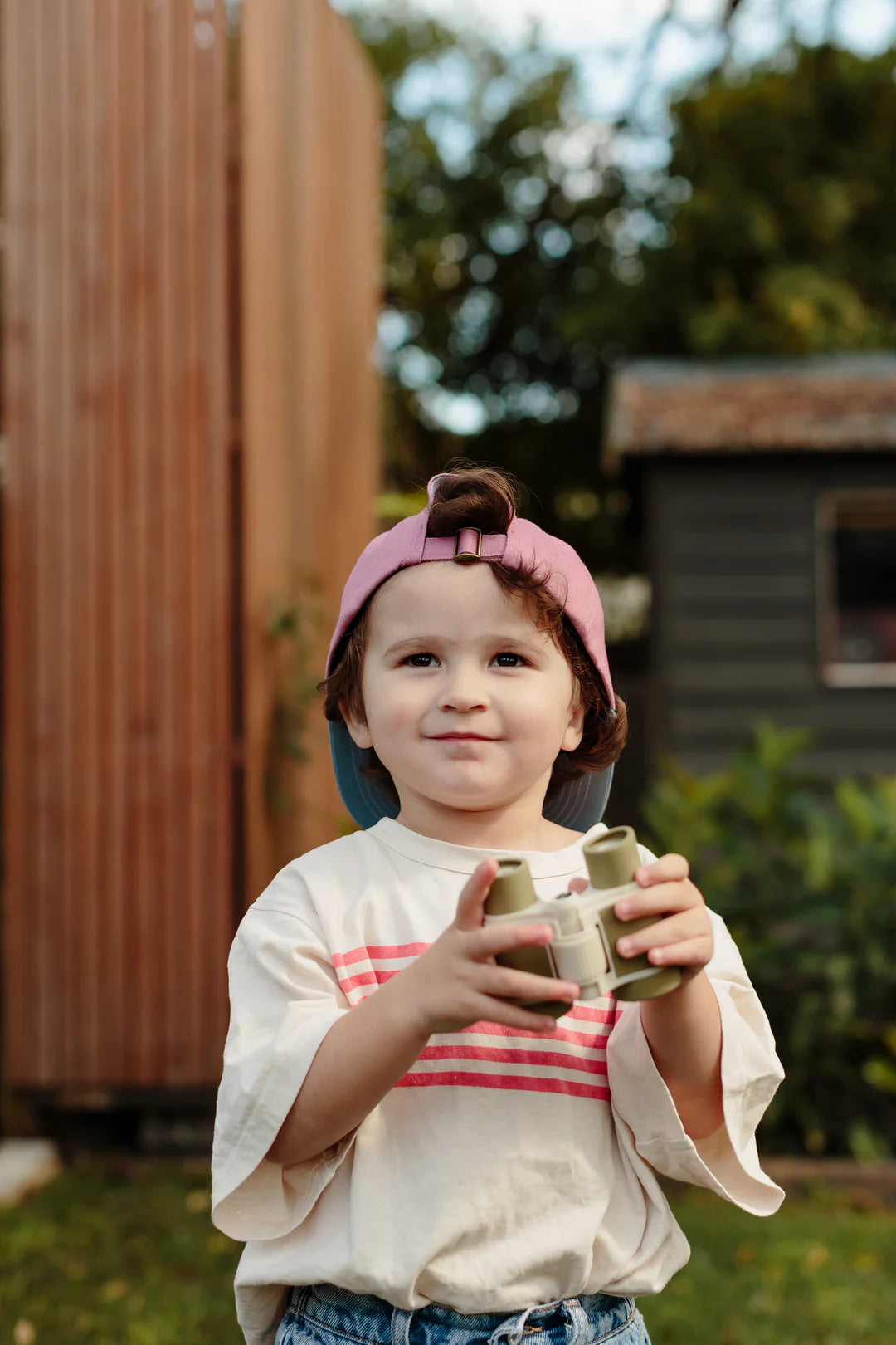 A young child with wavy brown hair, wearing a pink and blue cap backward, stands outside holding LITTLE DROP's Kids Explore Binoculars Moss with both hands. The child is dressed in a white shirt with pink stripes and blue jeans. A wooden structure and green foliage are visible in the background.