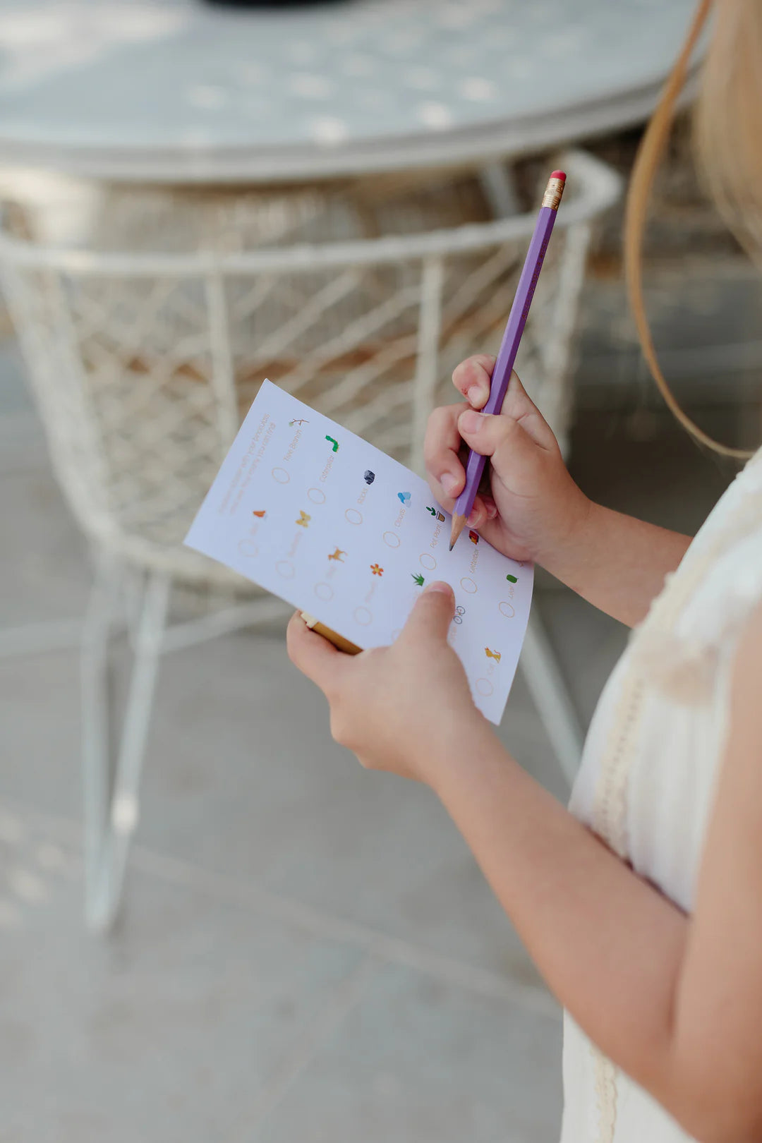 A person holding a sheet of paper and marking answers with a pencil. The paper contains multiple-choice questions with various small images and circles next to the options, resembling a scavenger hunt. In the background, there is a white wire chair and a table, beside which are placed LITTLE DROP's Kids Explore Binoculars Moss.