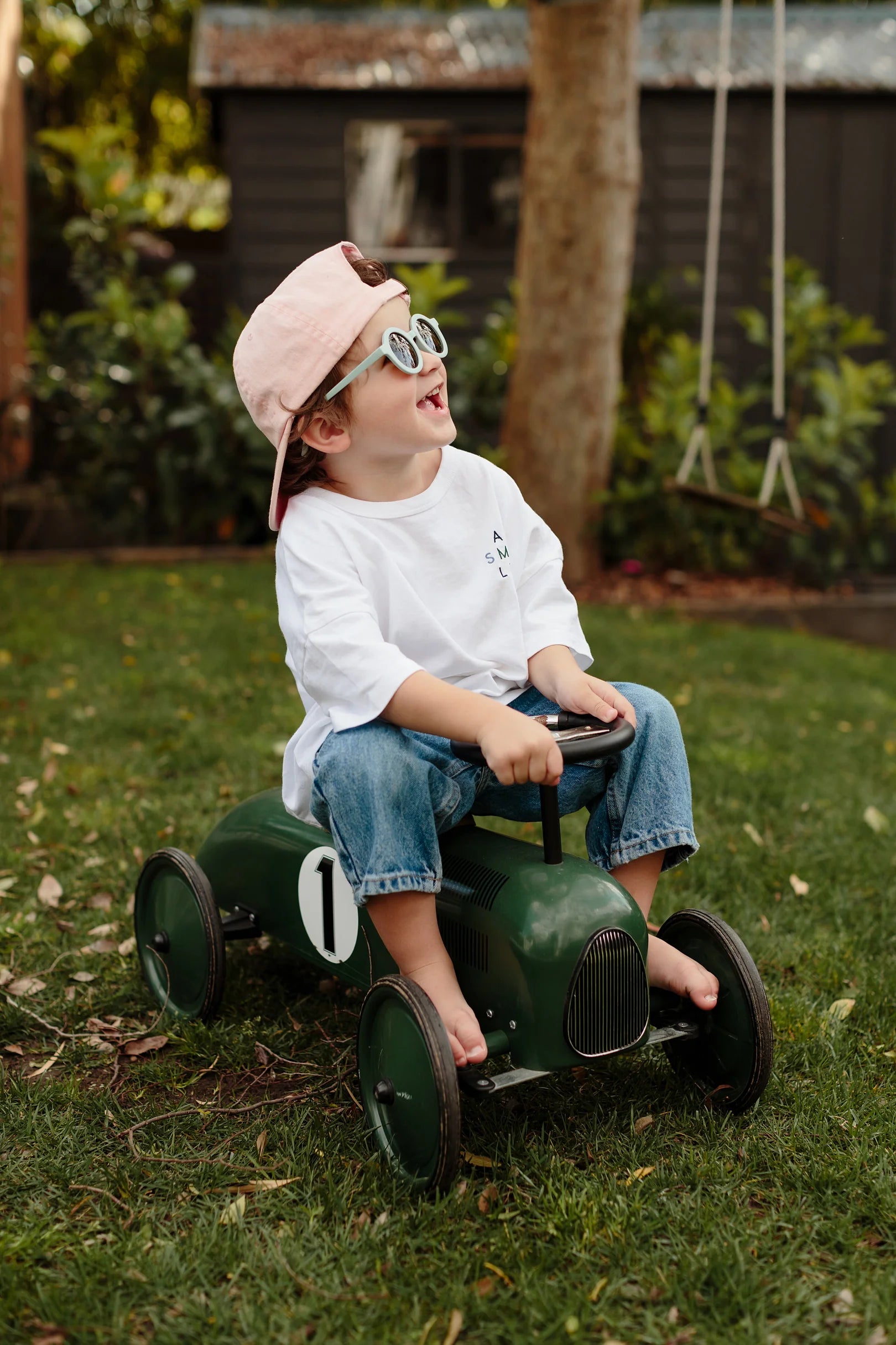 A young child sits on a small green toy car with the number one on its side, wearing a white shirt, rolled-up jeans, Kids Shades Sky by LITTLE DROP boasting UV400 protection, and a pink cap turned backwards. The child is smiling and looking upwards. A swing and a tree are visible in the background.